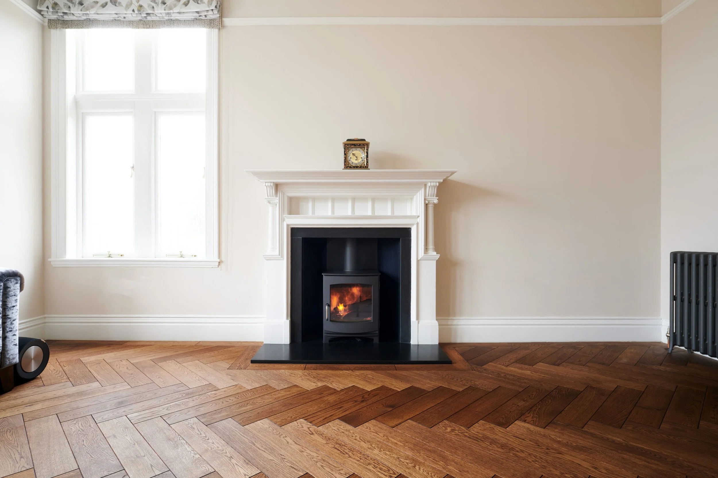 Living room with a fireplace and a clock on the mantle, large window to the left, hardwood floor, and light-colored walls.