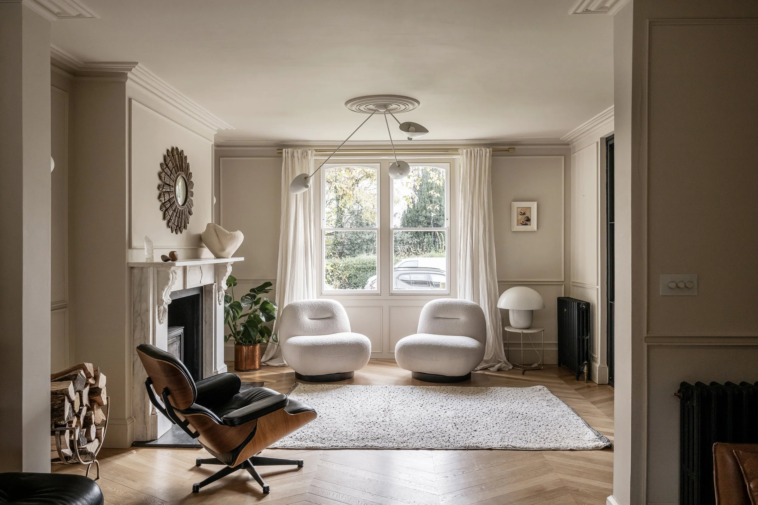A cozy living room with white walls, large window with curtains, two white armchairs, a small side table with a lamp, a fireplace with a decorated mantle, a potted plant, and a modern ceiling light fixture.