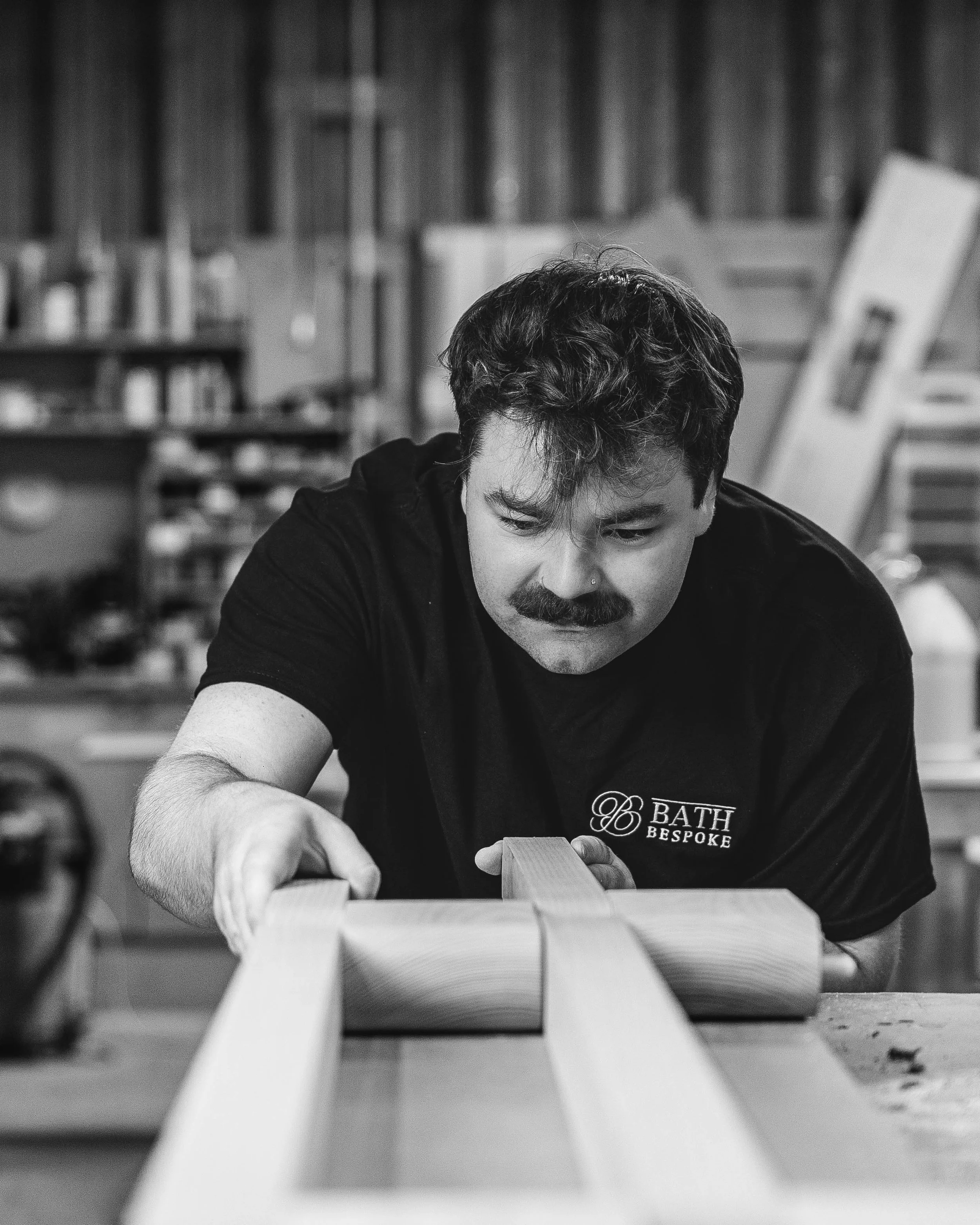 Black and white photo of a man with a mustache and curly hair working on a woodworking project in a workshop, wearing a shirt with a logo that reads 'Bath Bespoke.'