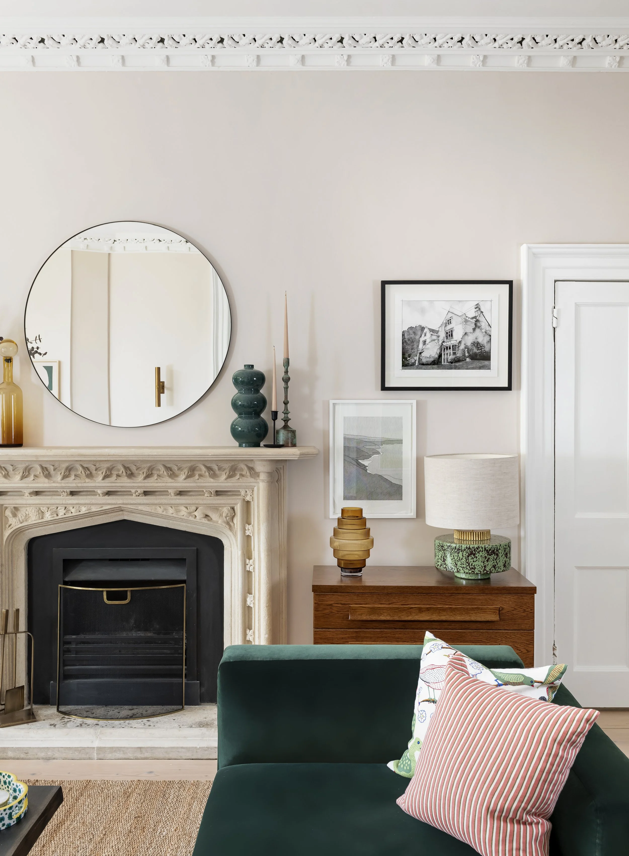 Living room with a fireplace, mirror, framed artwork, table lamp, decorative vases, and colorful pillows on a green sofa.