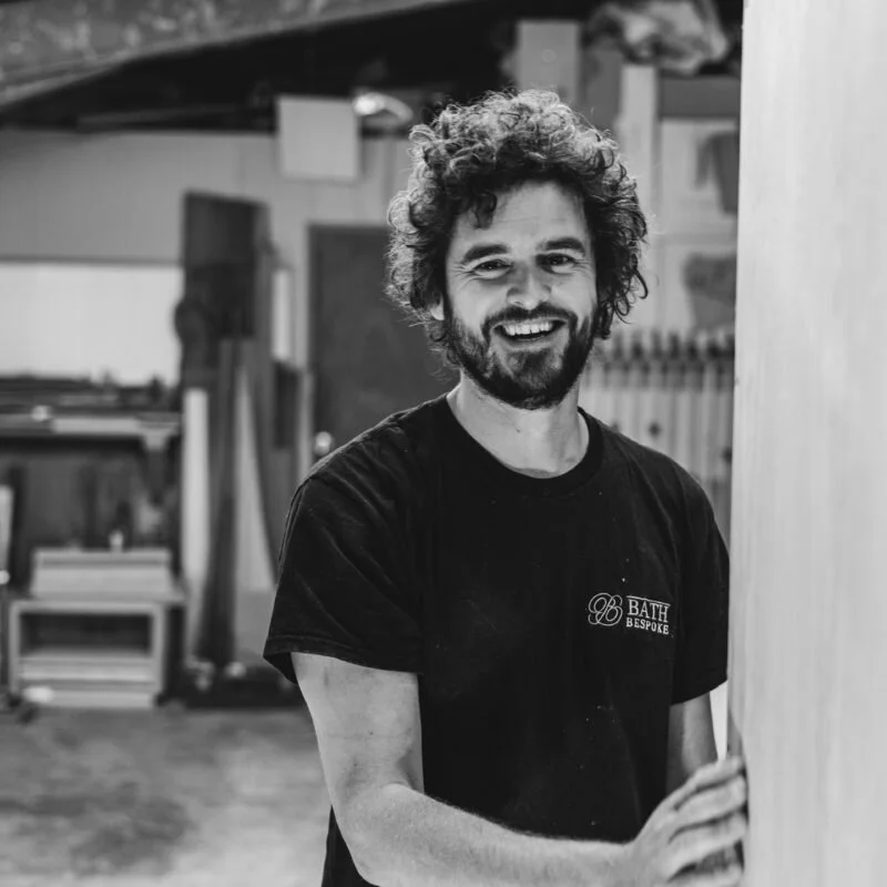 A smiling man with curly hair and a beard, wearing a black T-shirt, in a workshop or studio environment.