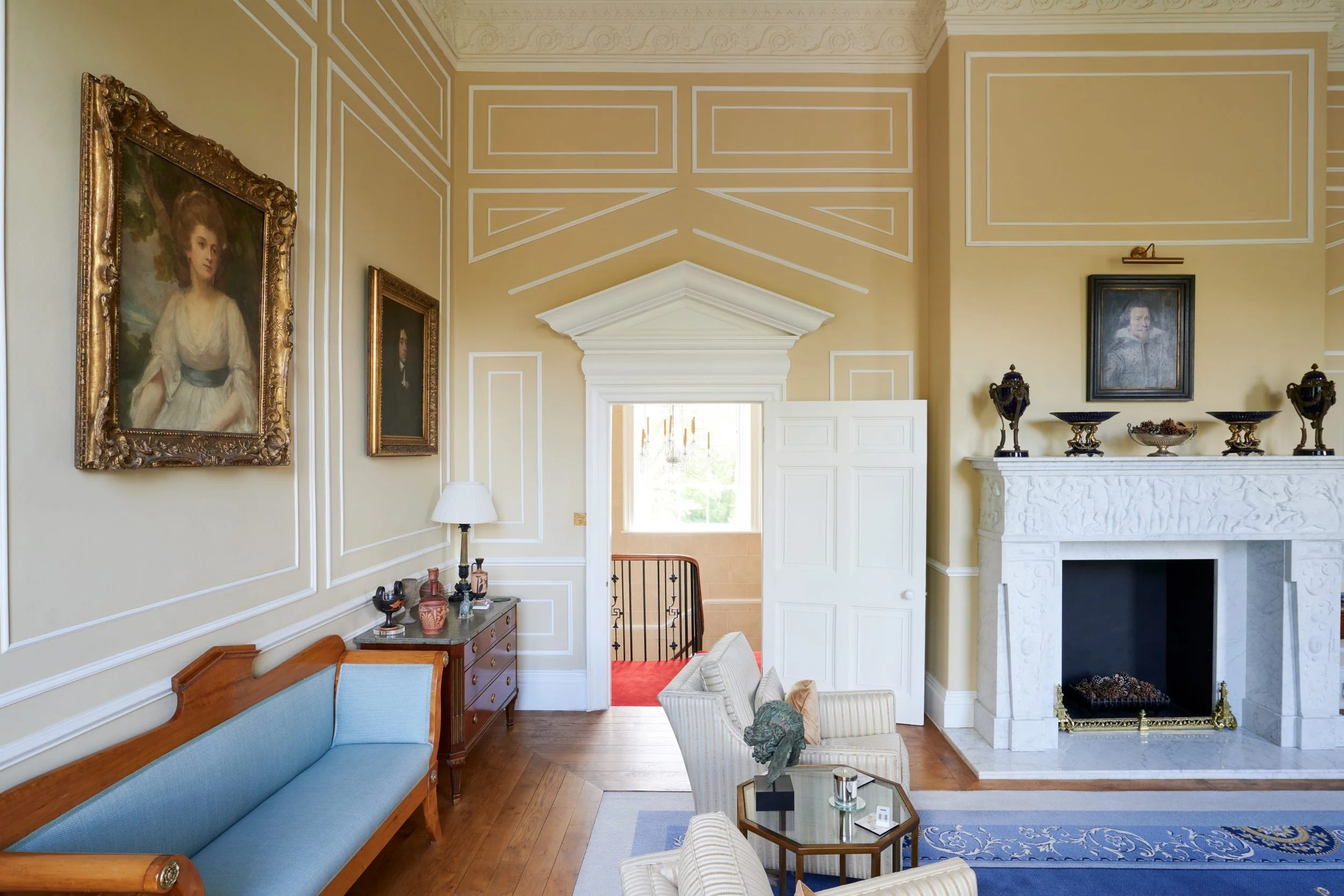 A spacious living room with ornate yellow walls and white trim, featuring artwork in gilded frames, a marble fireplace with decorative objects, and elegant furniture including a blue sofa and striped armchair.