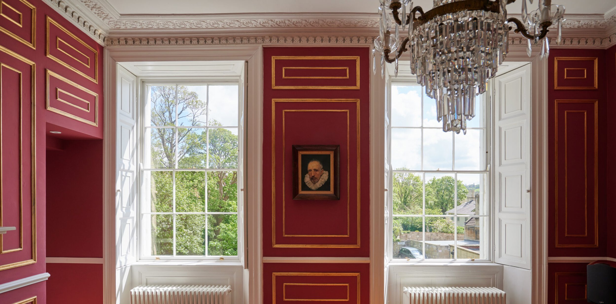 A bright room with large windows showing green trees outside. The walls are painted red with gold accents and white trim. There is a framed portrait of a man with a ruffled collar on the red wall. A crystal chandelier hangs from the ceiling.