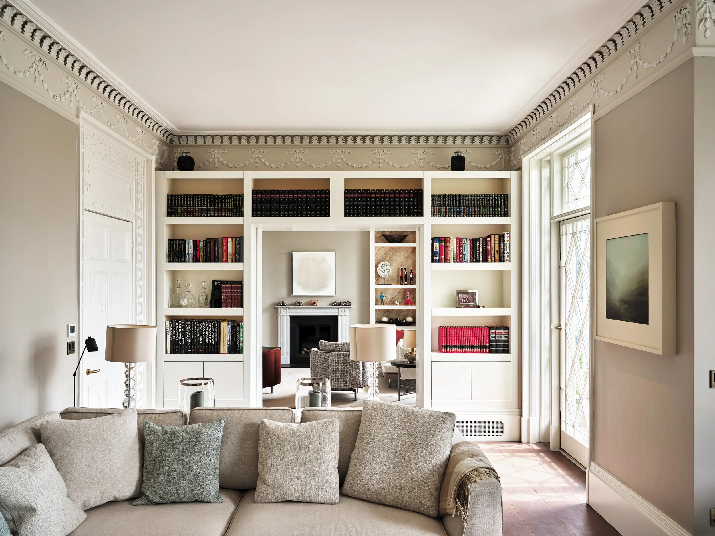 Living room with a beige sofa, cushions, a fireplace, and a white bookshelf filled with books, with a window on the right and ornate crown molding on the ceiling.