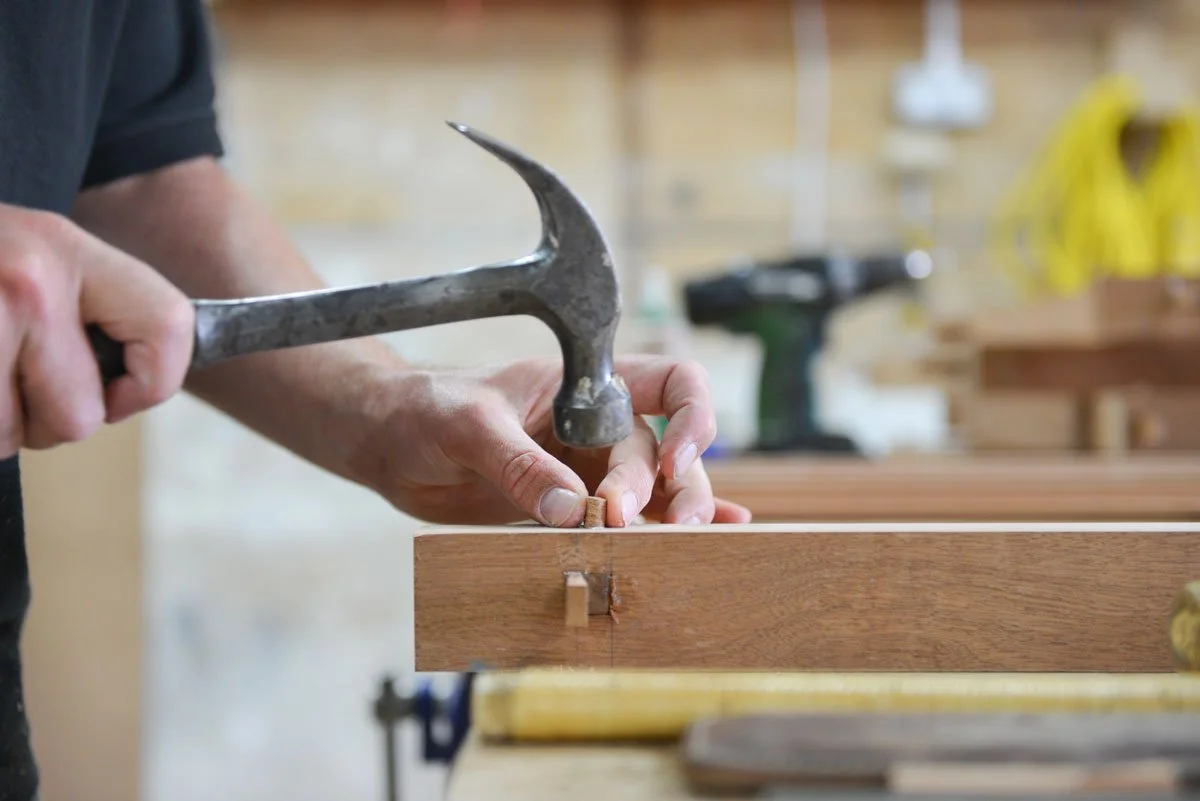 Person hammering a nail into a piece of wood in a woodworking shop.