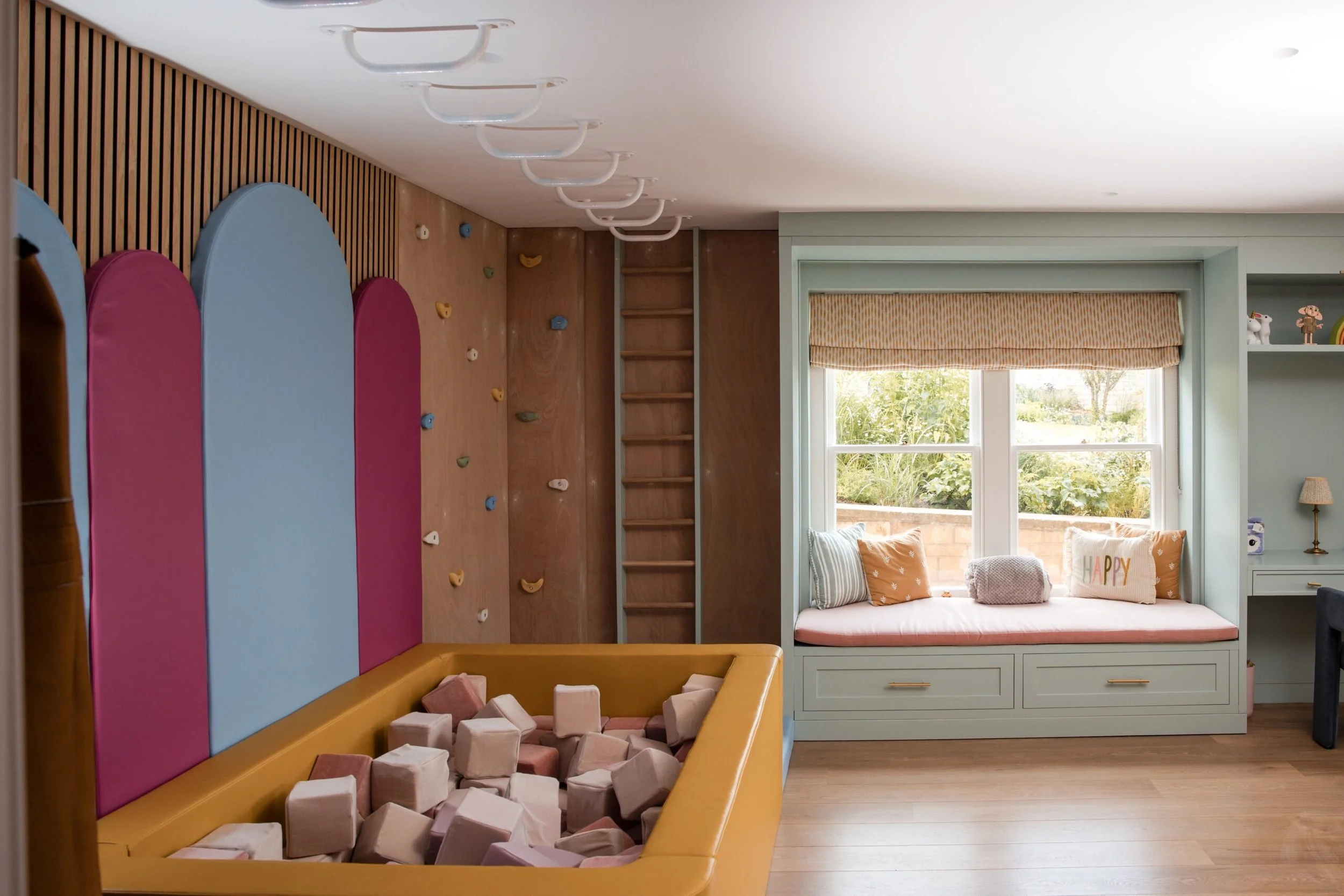 A children's playroom with a climbing wall, foam blocks, a window seat with pillows, and toys on a shelf, illuminated by natural light through large windows.