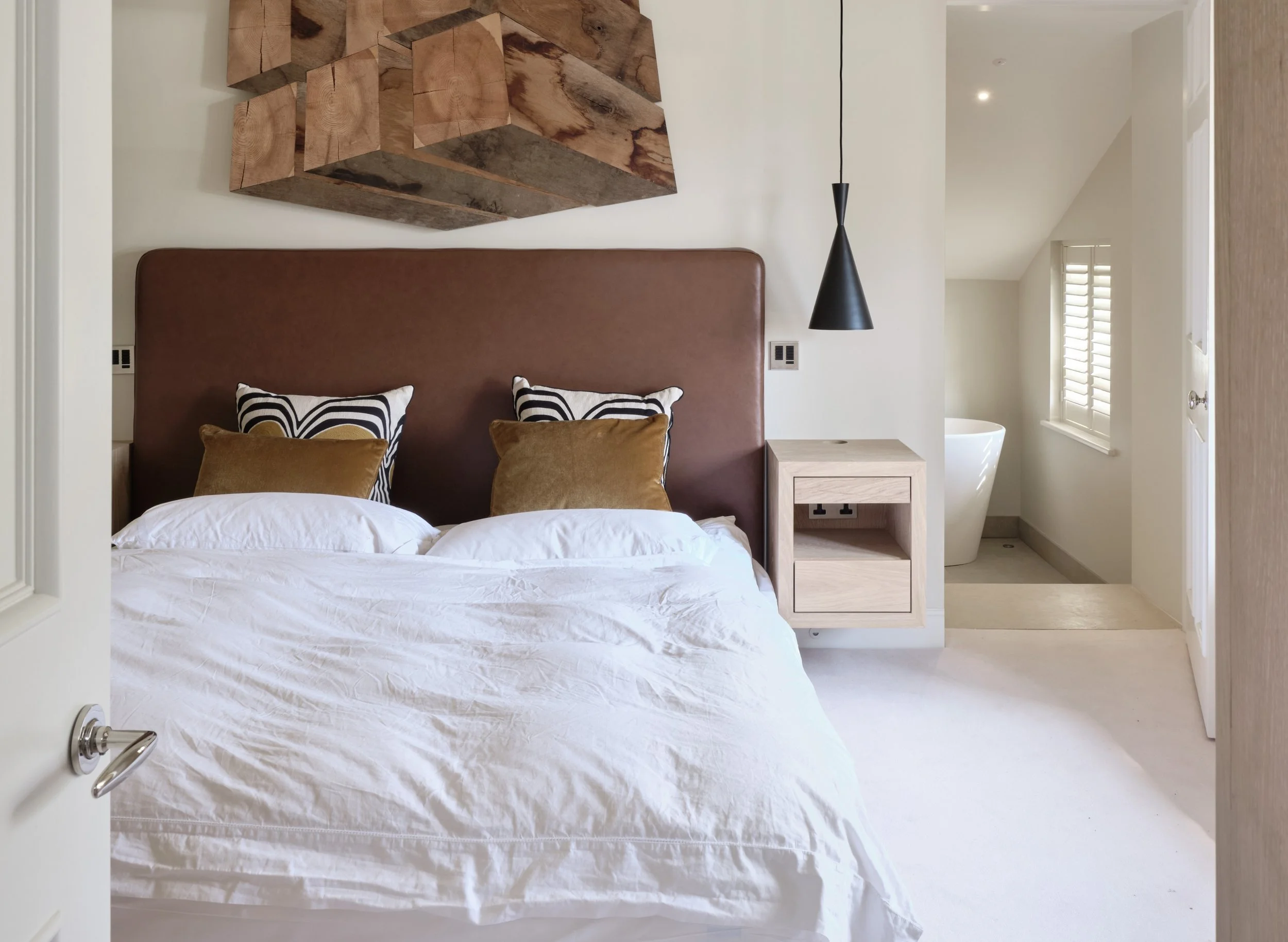 View of a modern bedroom with a bed, decorative pillows, wooden wall art, and a black pendant light hanging next to a nightstand. There is a window with shutters and a white chair in the background.