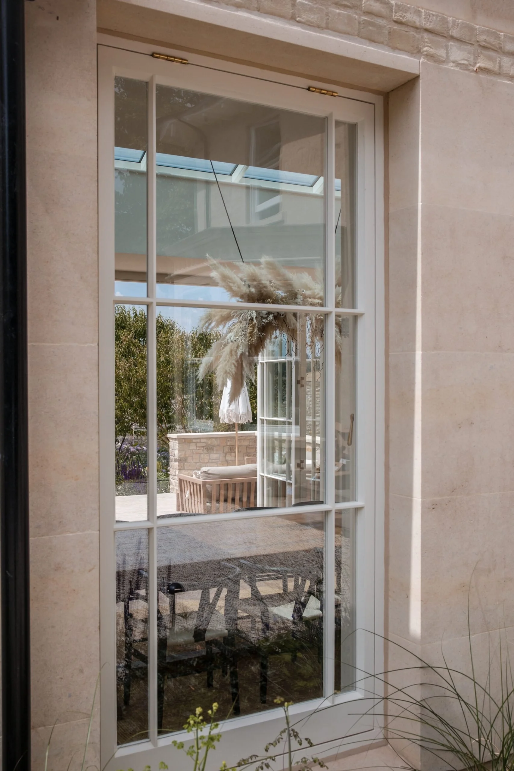 A large glass door with white framing, showing an outdoor patio with wooden furniture and a white umbrella, surrounded by greenery and potted plants.