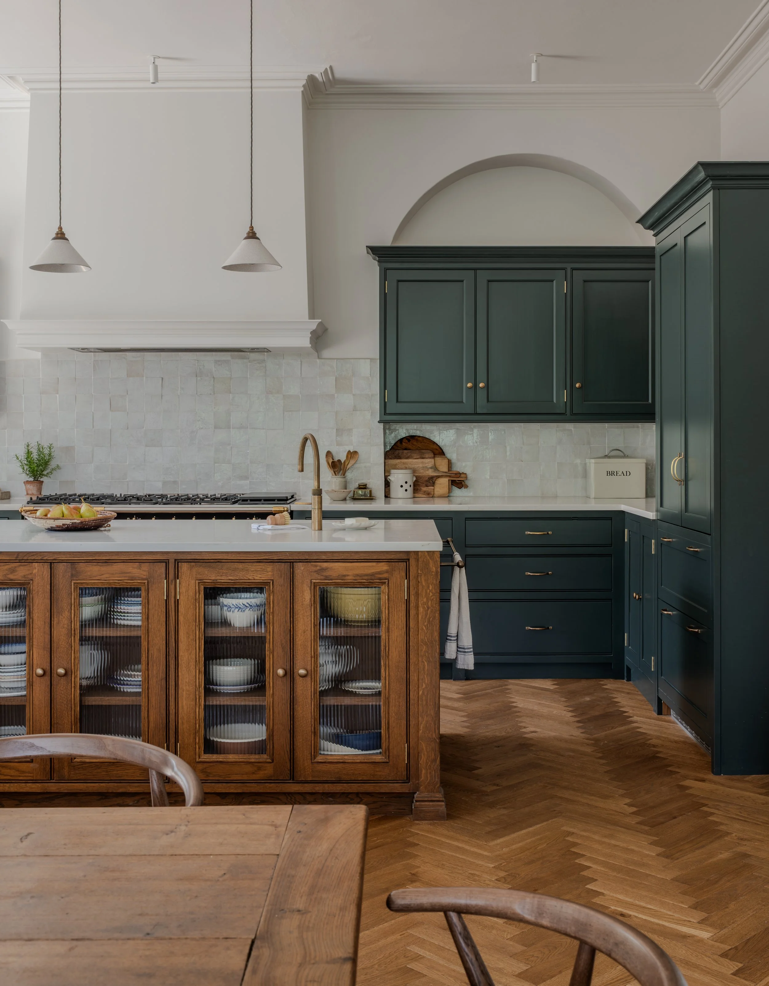 A kitchen with a wooden island and chairs, dark green cabinets, a white countertop, and a backsplash of light-colored tiles. The kitchen has pendant lights, a small potted plant, and decorative items on the counter.