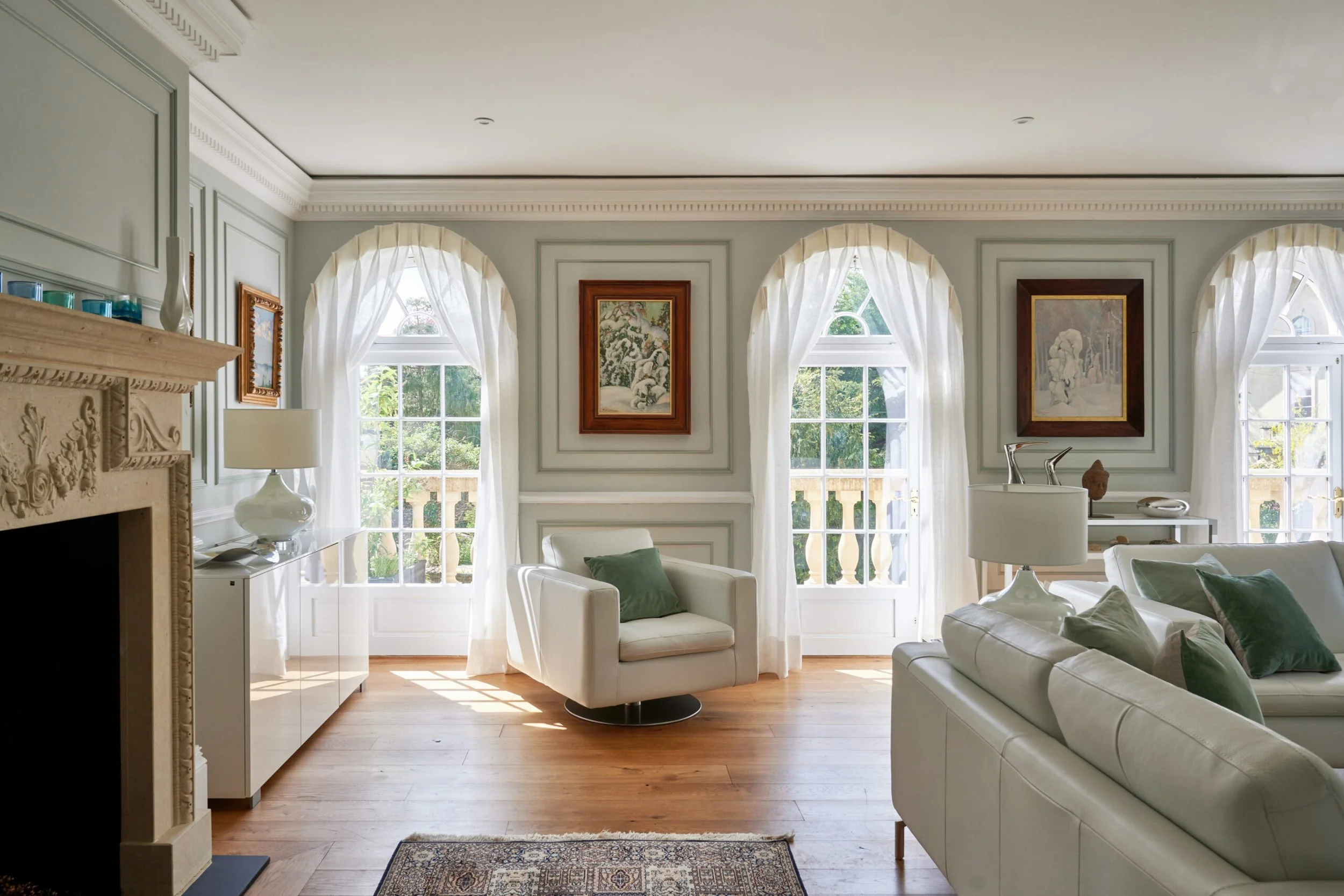 Bright living room with three arched windows dressed with white curtains, white sofas with green pillows, framed artwork on the wall, and wooden flooring.