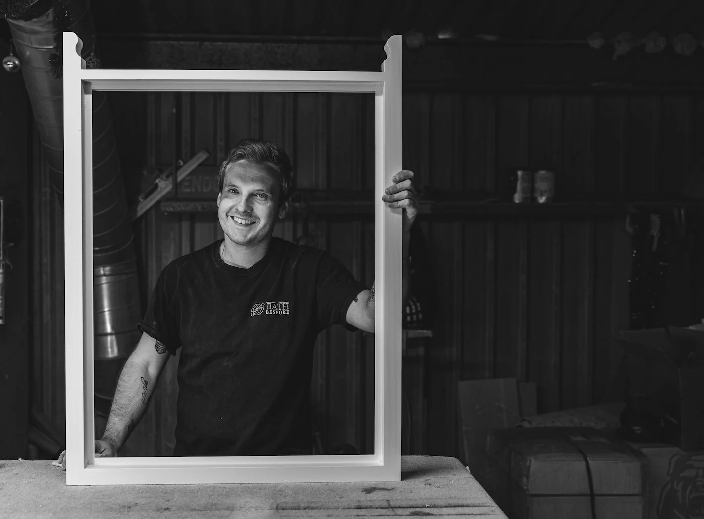 A smiling young man holding a picture frame with one hand, standing in a workshop or garage with wood paneling and various tools and objects in the background, in black and white.