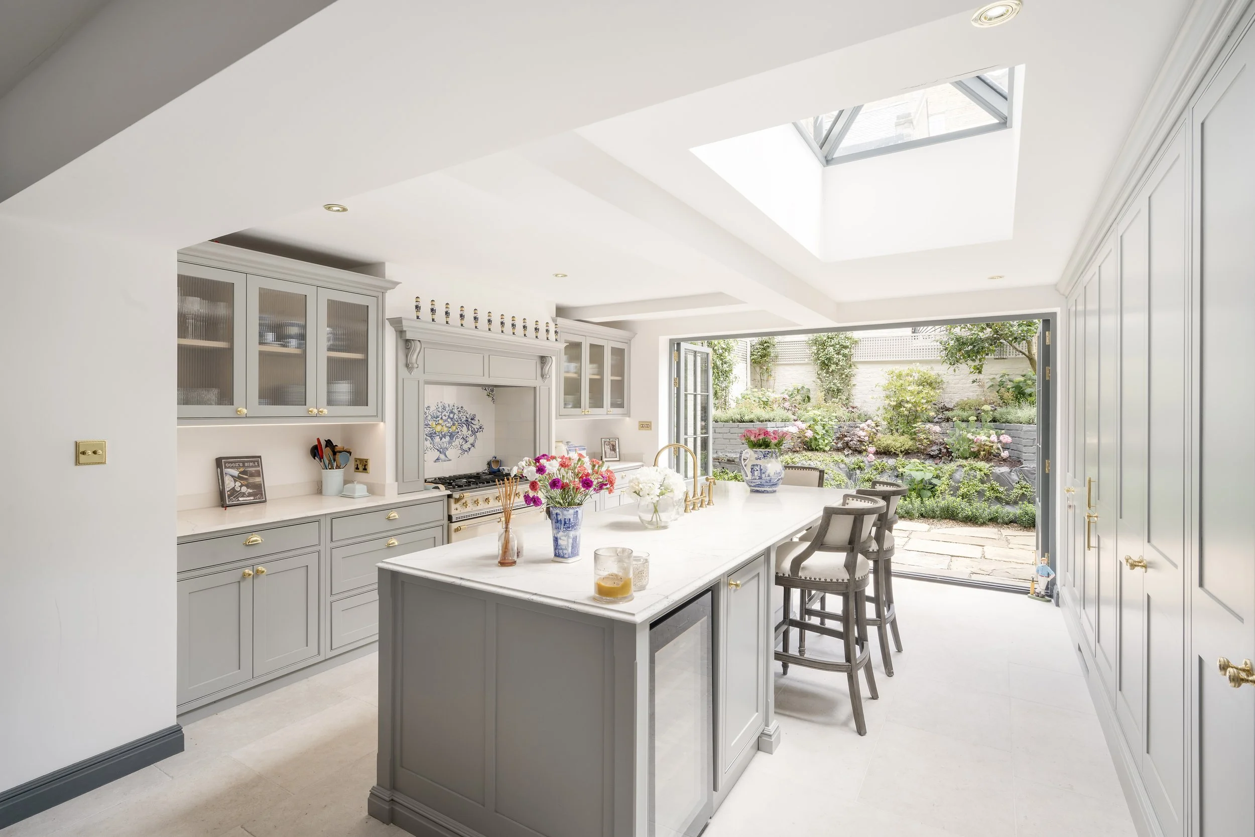 A bright and airy kitchen with white cabinets, a central island, and large glass doors opening to a garden with flowers and trees. Skylights let in natural light.