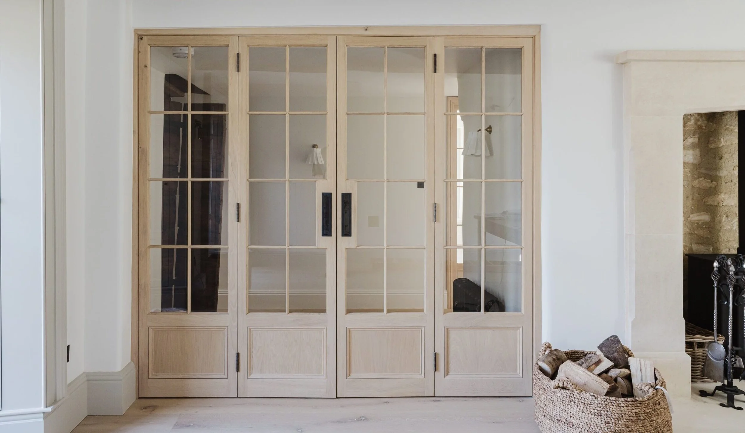 A set of wooden framed glass doors separating an interior room from another area, with a basket of firewood and a fireplace to the right.