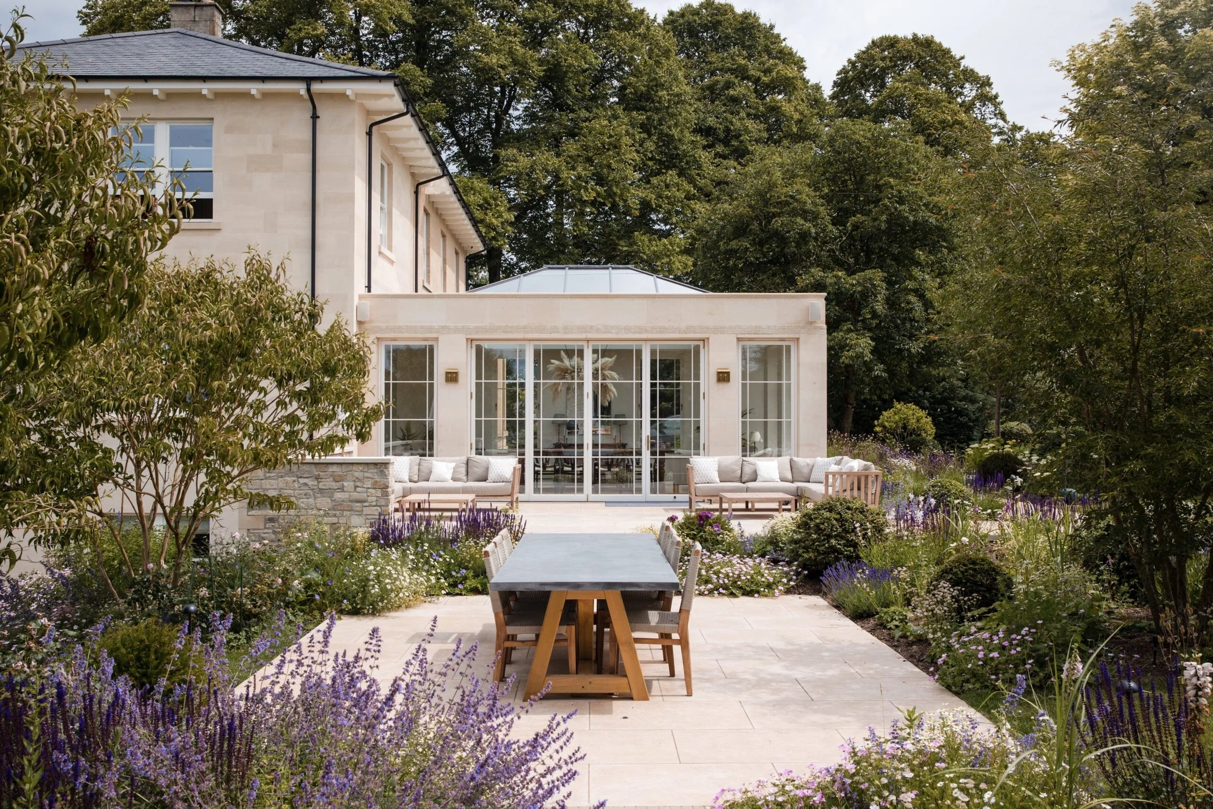 A backyard patio with a large table and chairs, surrounded by a garden with purple and white flowers, trees, and a white house with large glass doors.