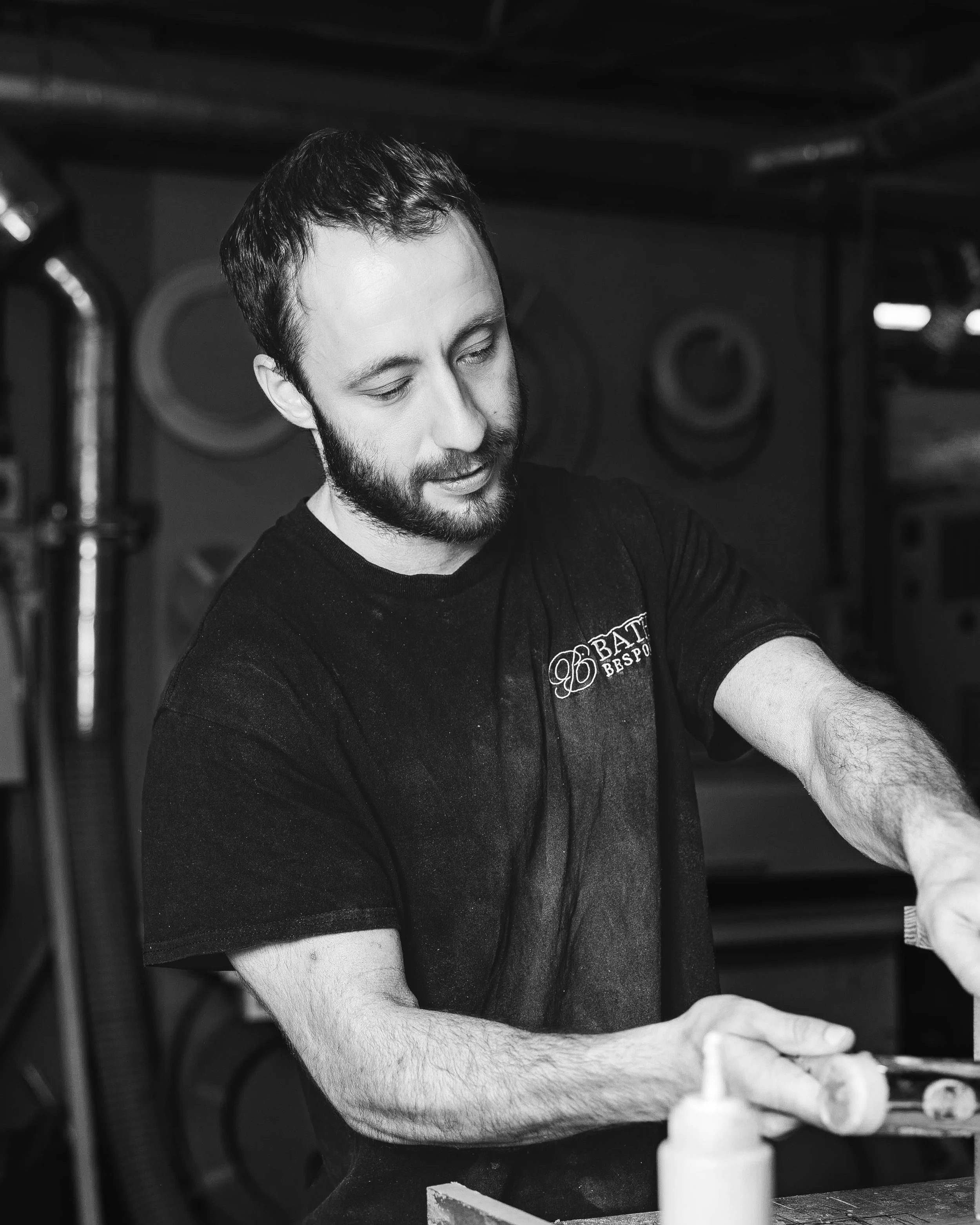 A man with a beard and short hair, wearing a black T-shirt, is focused on an activity at a counter. The background includes industrial elements like pipes and circular designs, and a bottle of glue or sauce is visible in the foreground.
