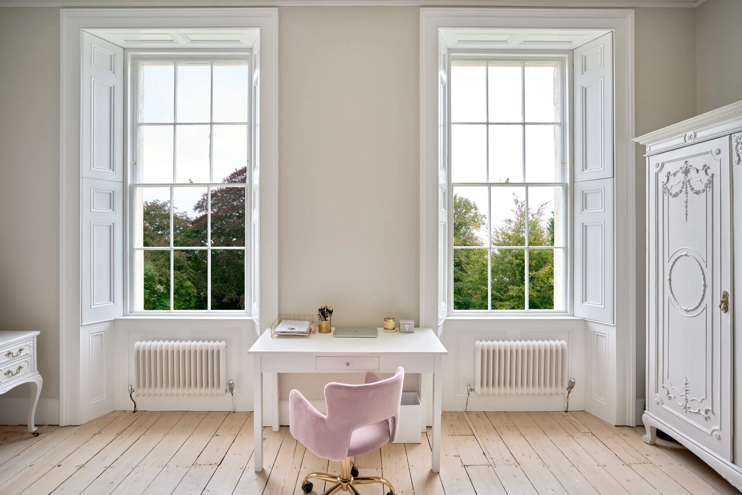A bright space with two large windows featuring white trim and shutters, overlooking trees outside. A white desk is positioned beneath the windows with a pink velvet office chair in front. On the desk are a closed laptop, a gold cup with pens, a notebook, and a candle. The room has light wooden flooring, white walls, and a vintage white cabinet with ornate detailing on the right.
