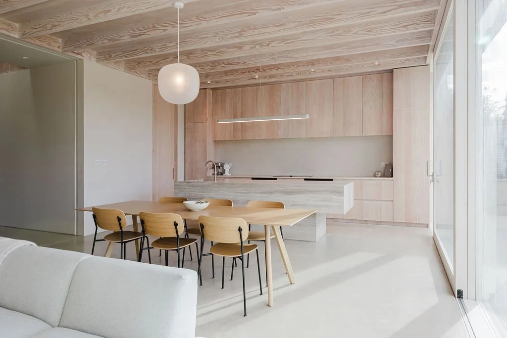 Modern kitchen and dining area with wooden cabinetry, a light wooden dining table, six chairs, and a large window or sliding glass door letting in natural light.