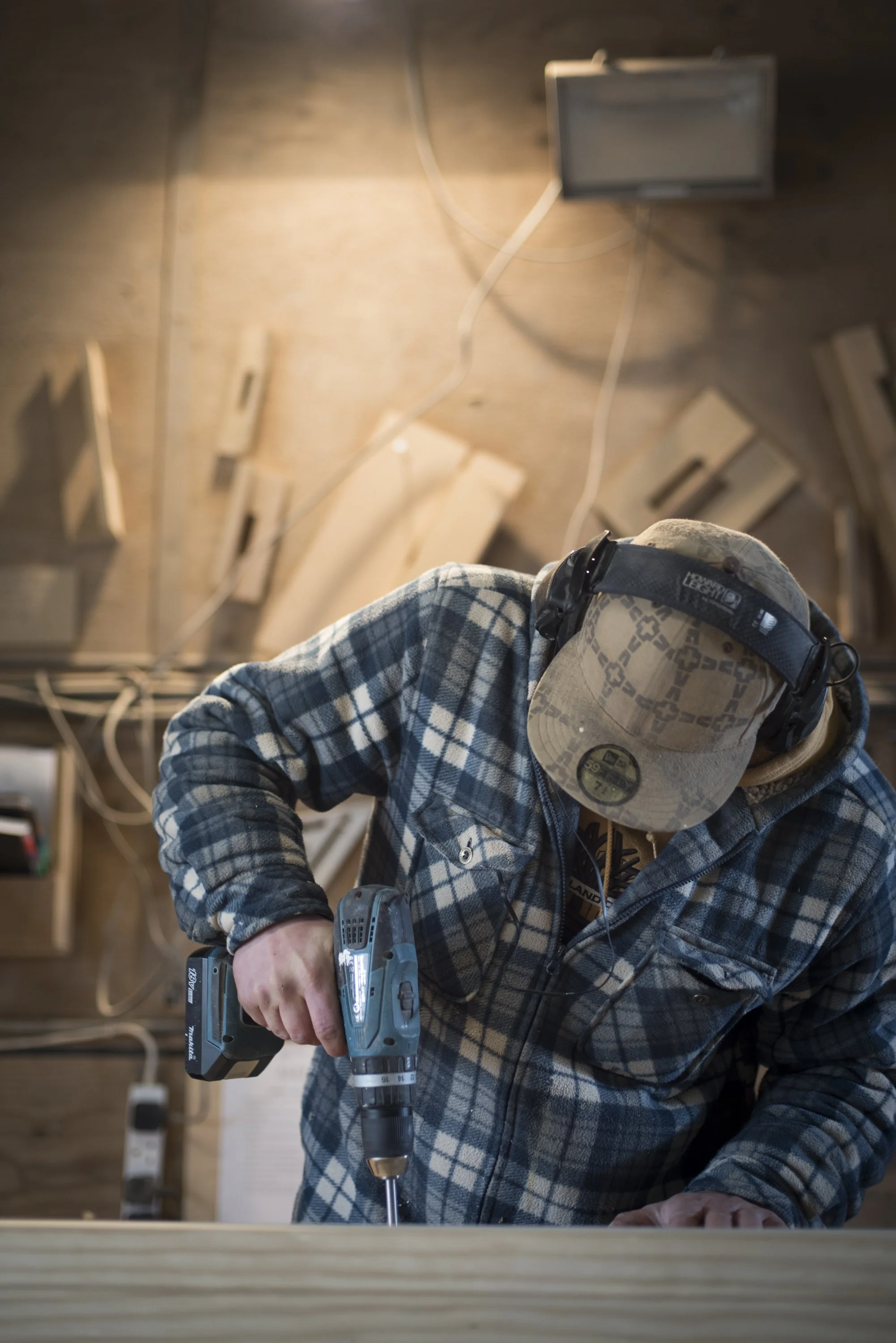 A person in a plaid shirt and a cap using a cordless power drill on a piece of wood in a woodworking or construction setting.