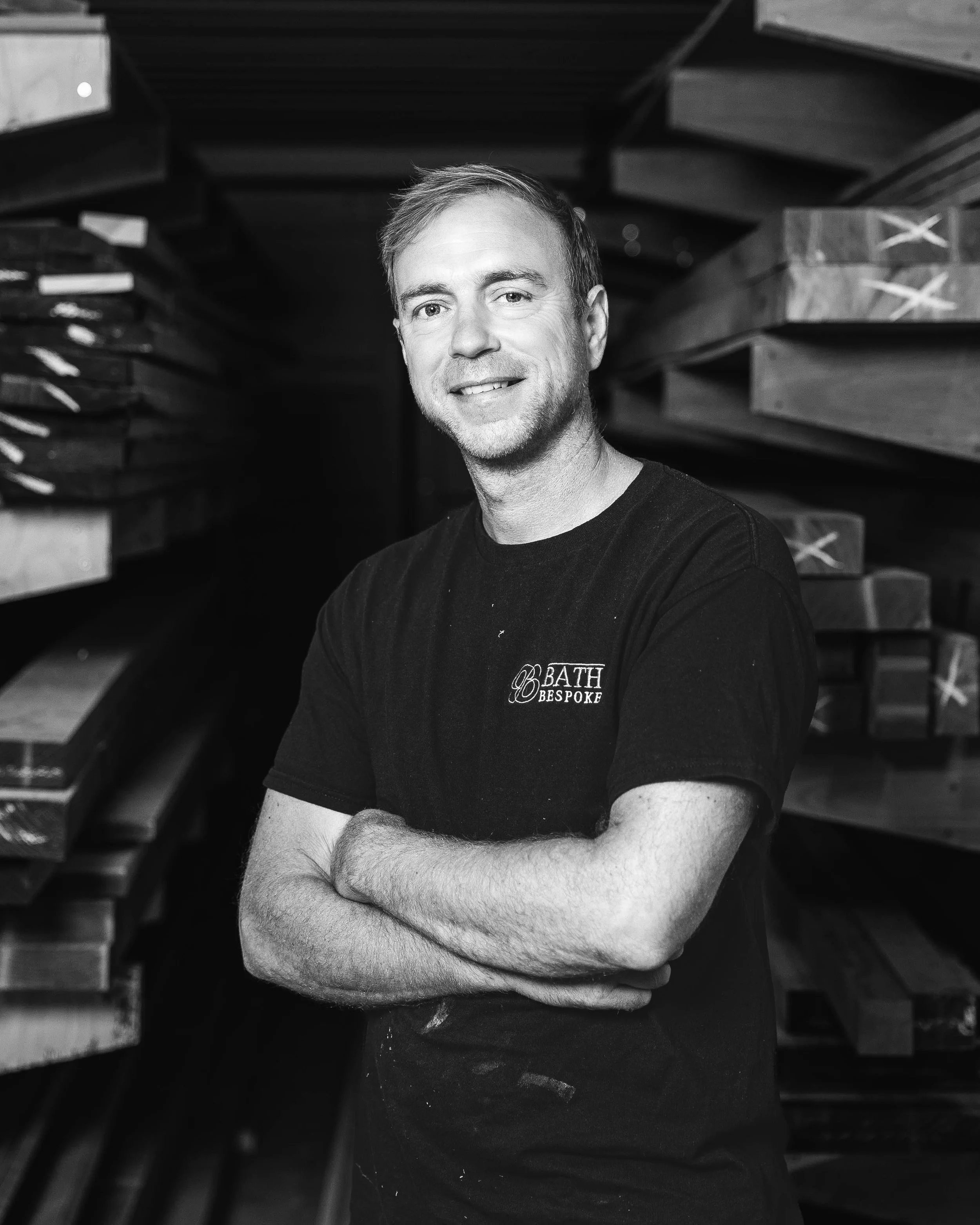 Black and white photo of a man standing with arms crossed in a woodworking shop, surrounded by stacks of wooden planks and materials.