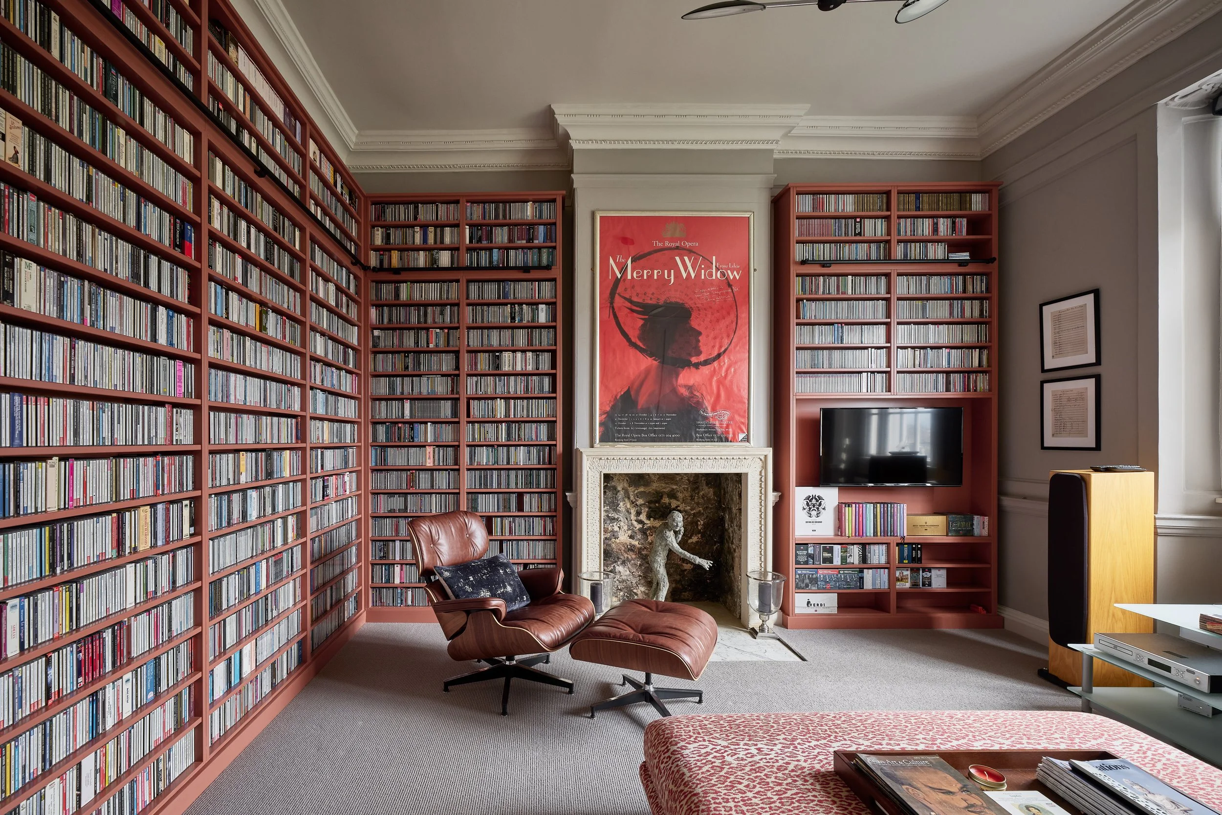 A living room with tall red bookshelves filled with books and CDs, a fireplace with a sculpture of a woman, a leather lounge chair with a pillow, a leopard-print ottoman, a small side table with magazines, framed artwork on the wall, a television, an