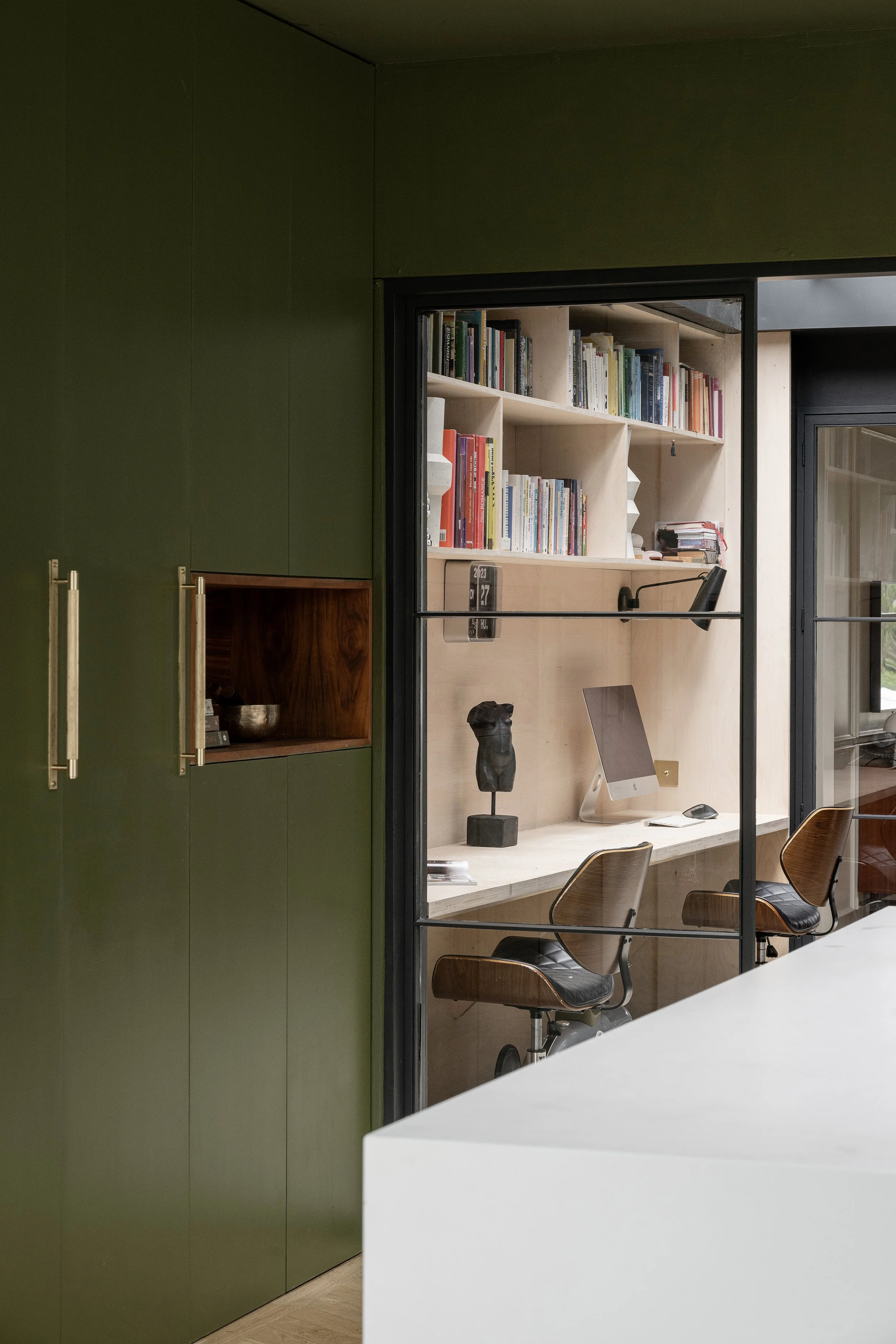 A modern office space with a white desk and wooden chairs. Behind the desk is a built-in wooden shelf with a bust sculpture on a stand, a computer monitor, and a window with a glass wall revealing a bookshelf filled with various books.