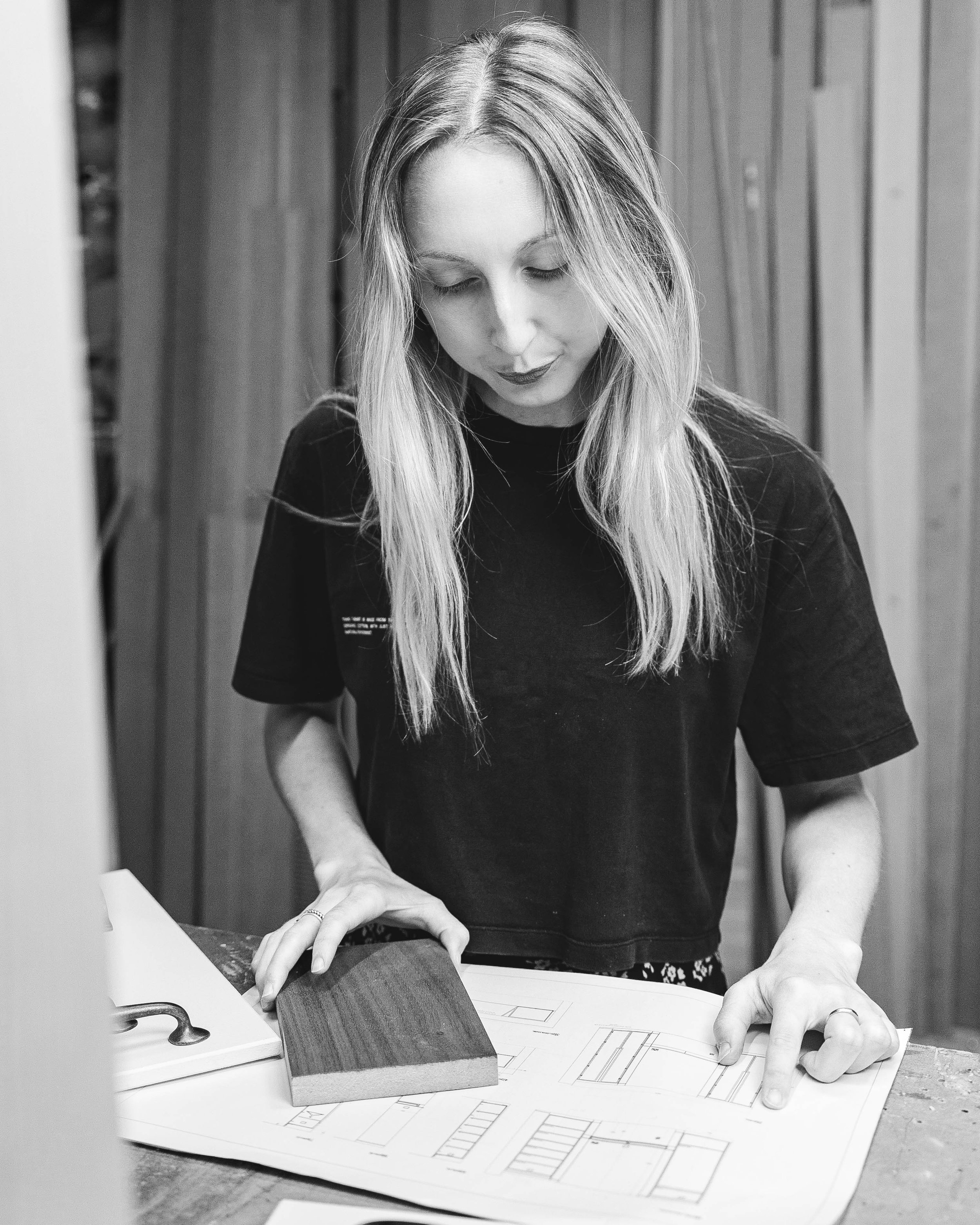 A woman with long blonde hair, looking down and pointing at architectural plans on a table, with a wooden sample and a clamp nearby, in a room with wooden panel walls.