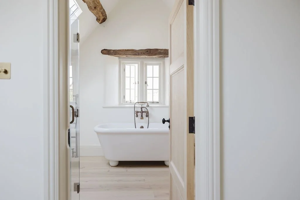 Bathroom with a white clawfoot bathtub under a window with two small panes, exposed wooden beam on ceiling, light-colored walls, and hardwood floor.