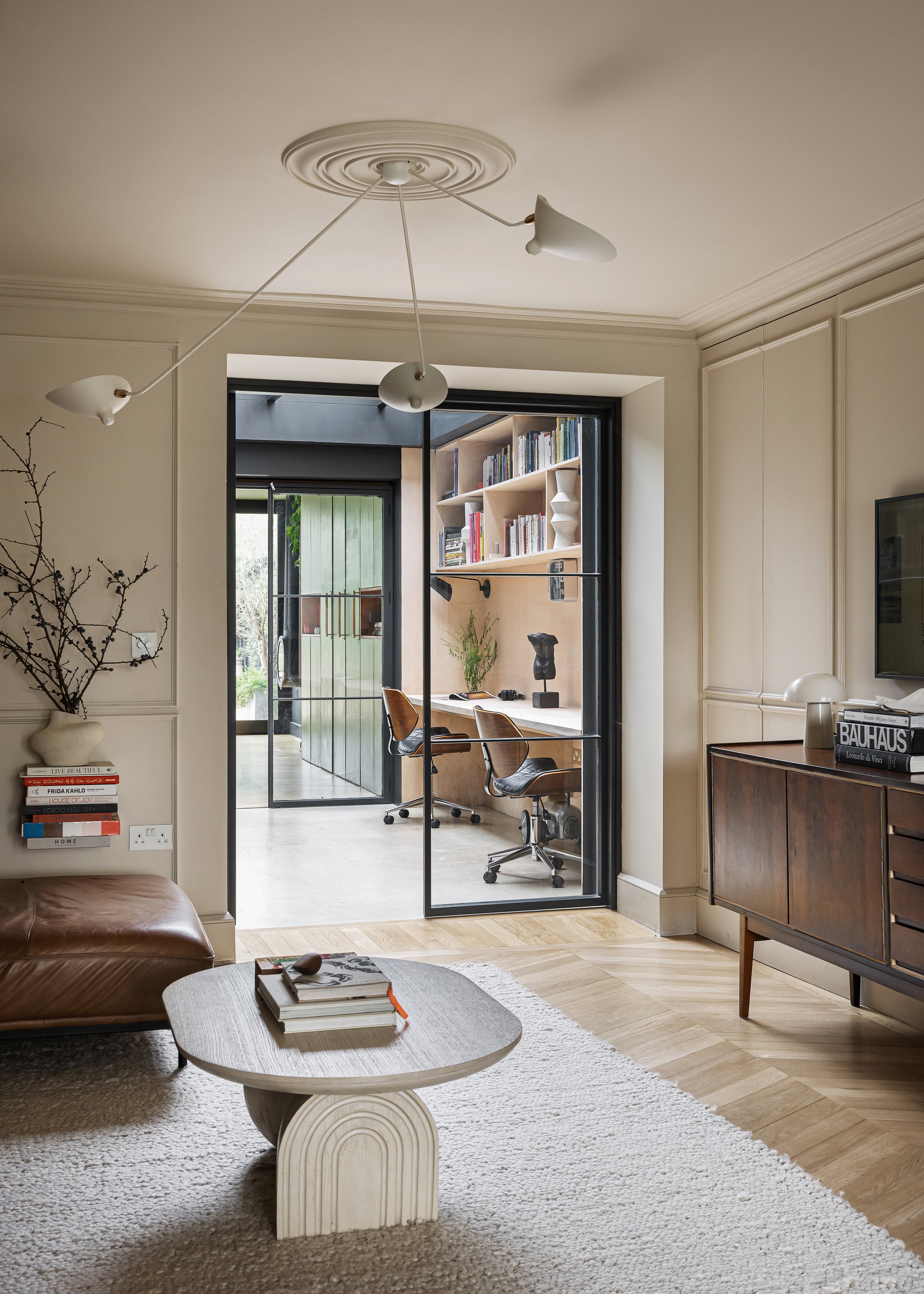 Living room with a leather sofa, wooden coffee table with books, a white shag rug, and an ornate ceiling light fixture. Through black-framed glass doors, a home office area with a desk, rolling chairs, and a bookshelf visible in the background.