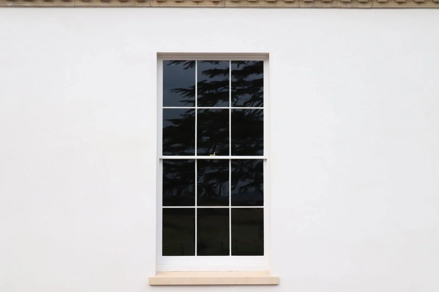 A white wall with a large window divided into twelve panes, reflecting a partly cloudy sky and trees.