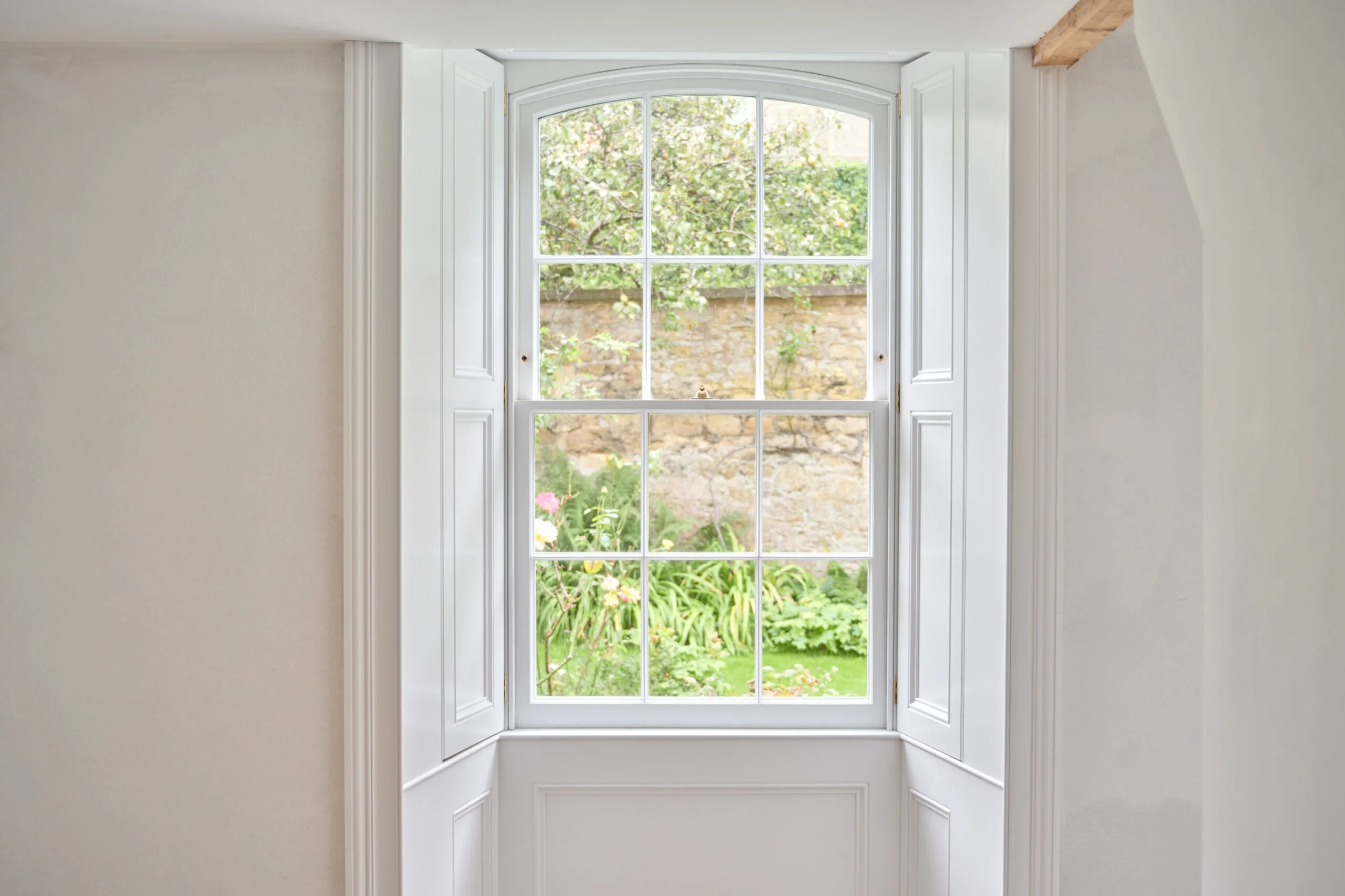 A large white-framed window with open white shutters shows a lush garden outside with green plants and a stone wall.