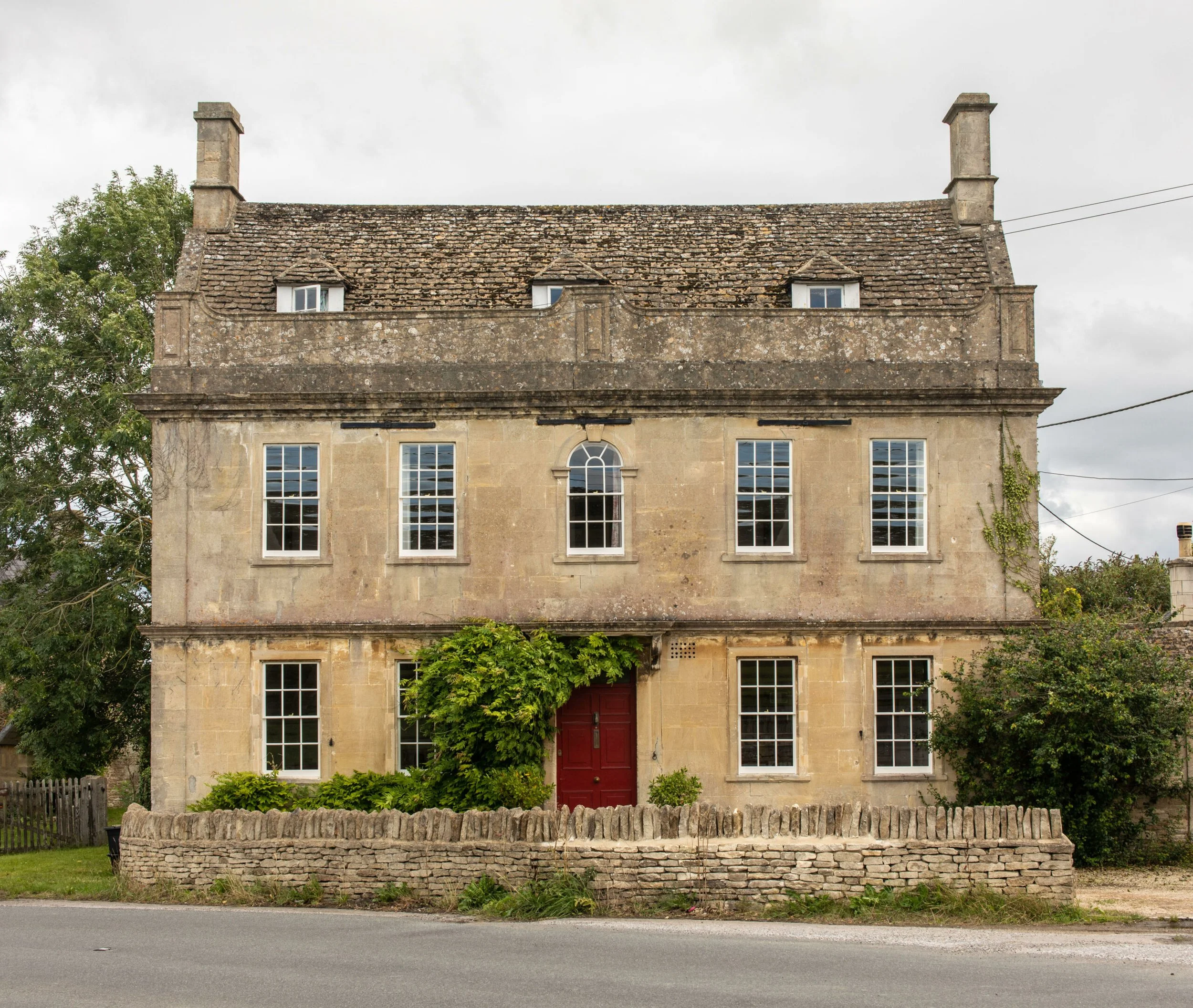 A large, old stone building with a peaked roof covered in weathered shingles. The building has several windows, a large red front door, and is surrounded by greenery and a low stone wall.