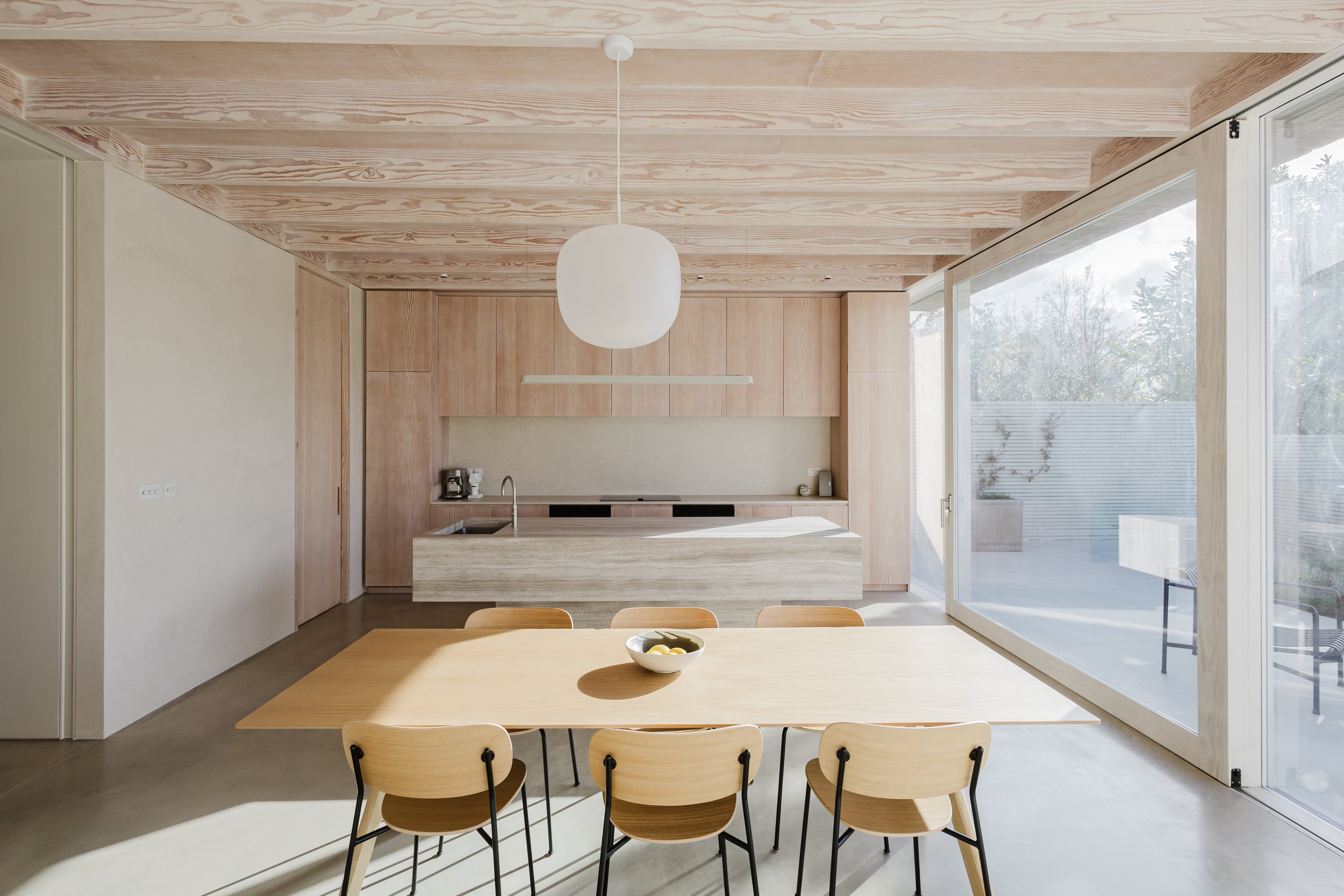 Modern kitchen with natural wood accents and large sliding glass doors leading to outdoor space, featuring a wooden dining table with chairs and a minimalistic pendant light.