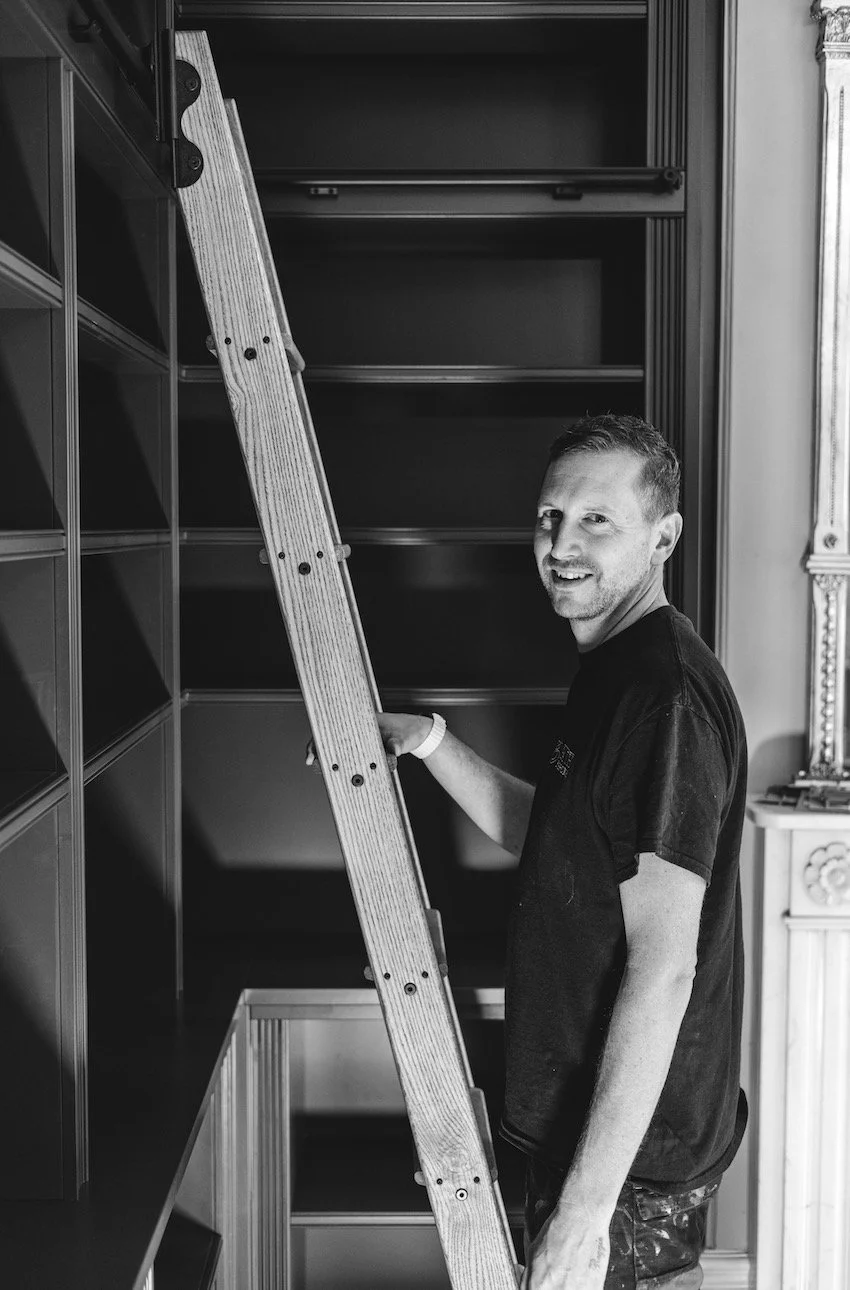 A man standing next to a ladder in front of empty bookshelves, smiling at the camera.