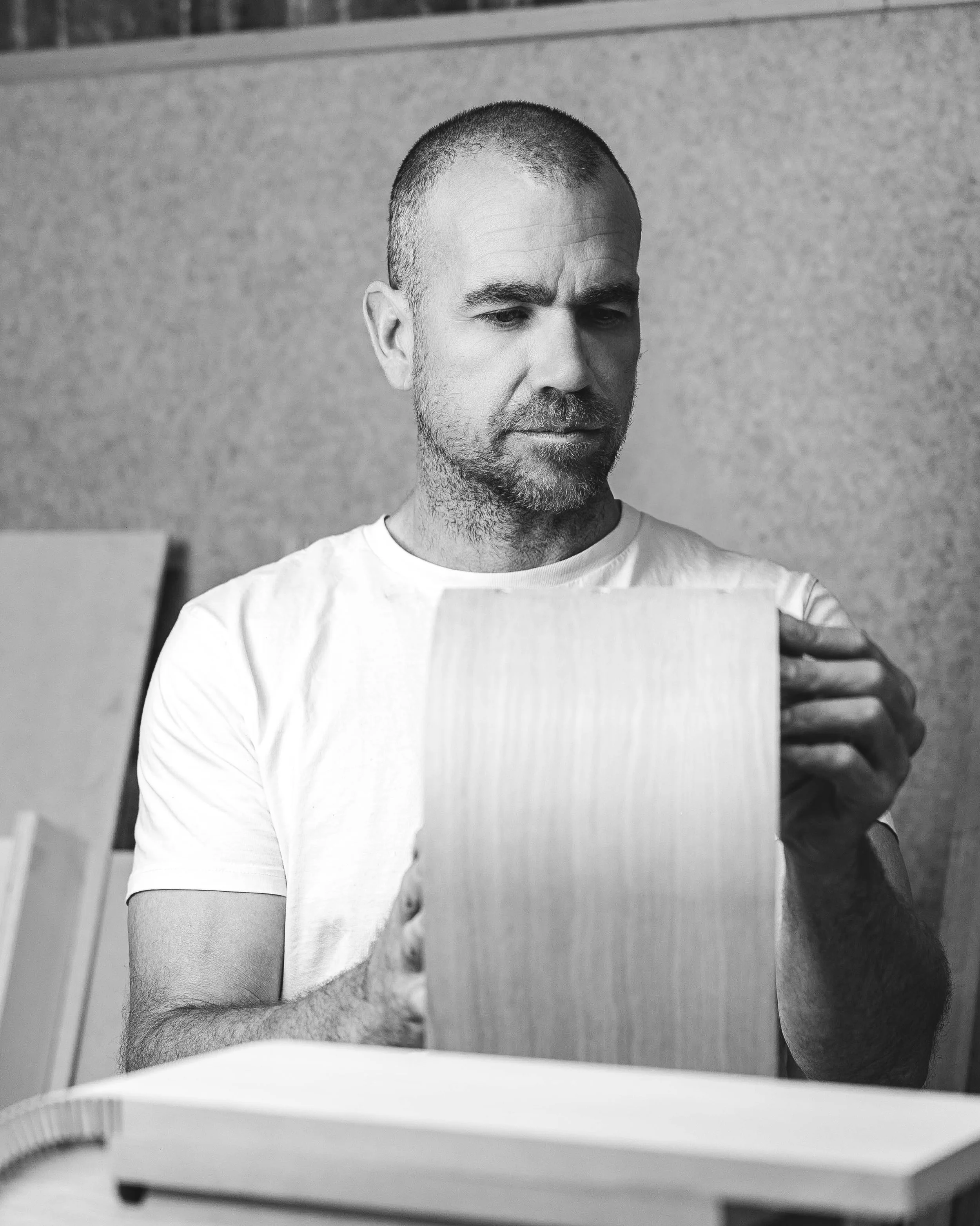 Black and white photo of a man with short hair and a beard, wearing a white t-shirt, holding a piece of paper or card in front of him, with a plain wall background.