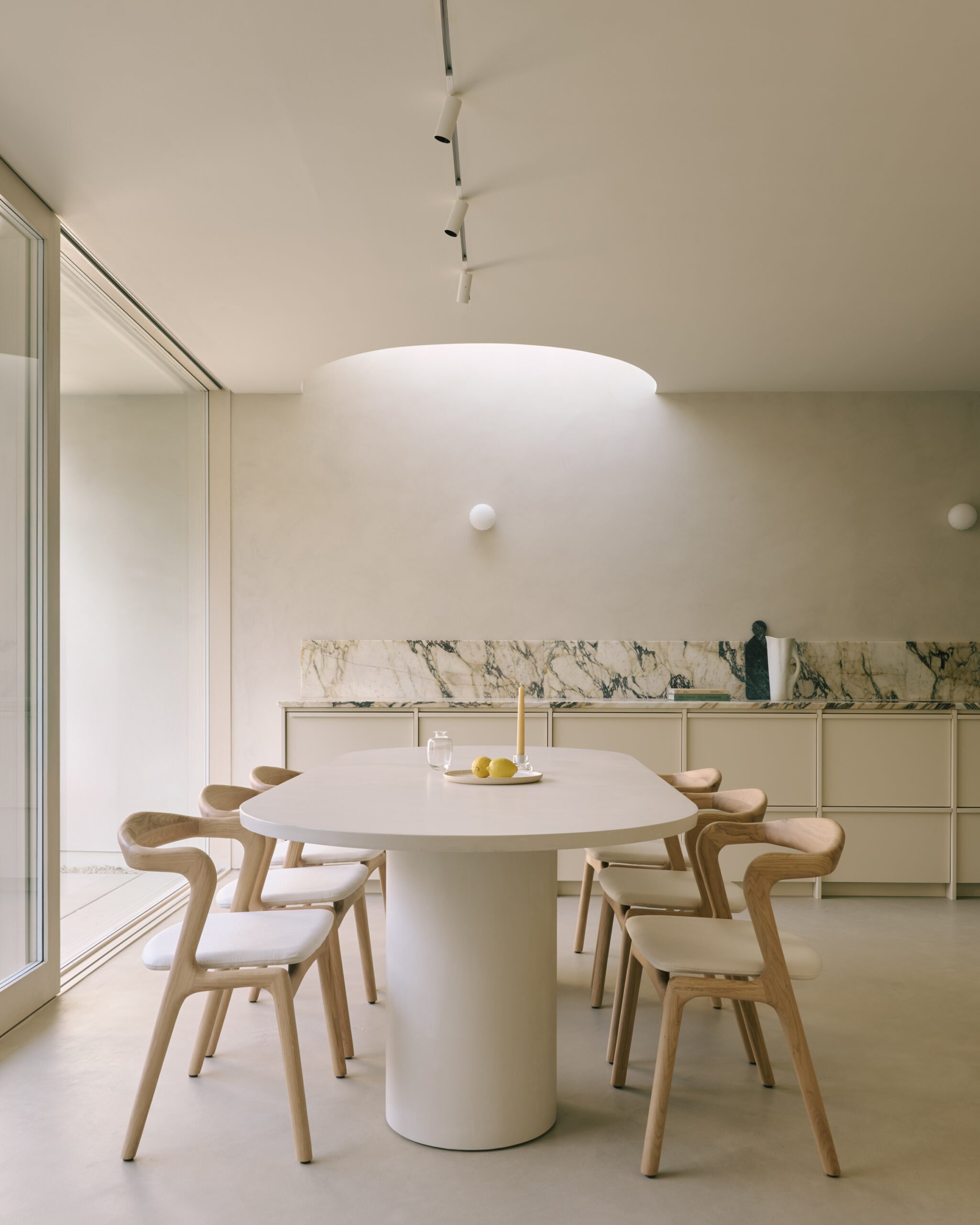 A modern dining area with a large white round table surrounded by seven wooden chairs with light-colored cushions. On the table, there is a white plate with three yellow lemons, a white candle, and a small glass. The background features a marble backsplash, a white cabinet, and a minimalist decor. The space is well-lit by natural light coming from a large glass door or window to the left.