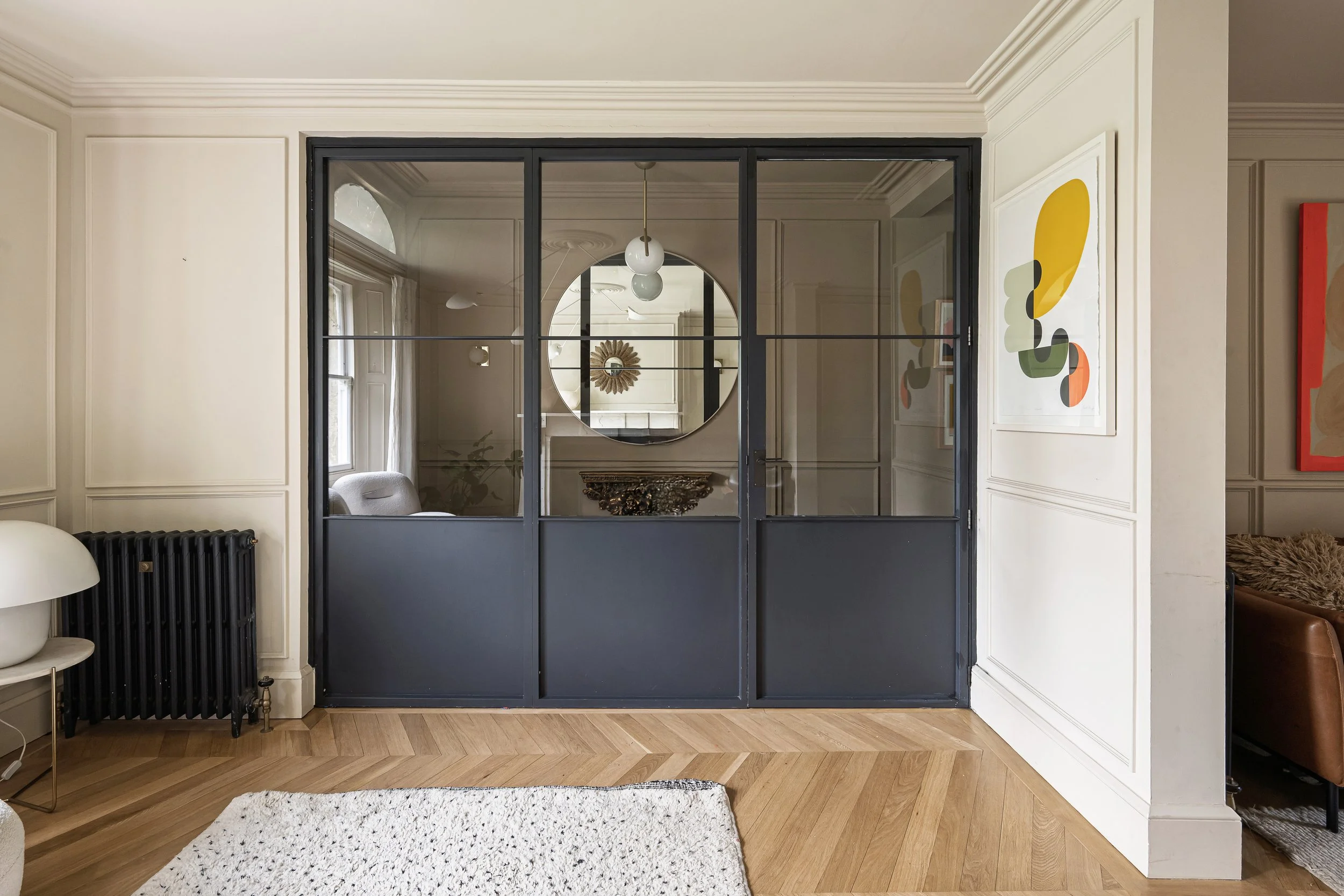 Interior view of a room with a black-framed glass partition, white walls with decorative paneling, a large round mirror, modern art on the walls, hardwood floor, and a white shag rug.