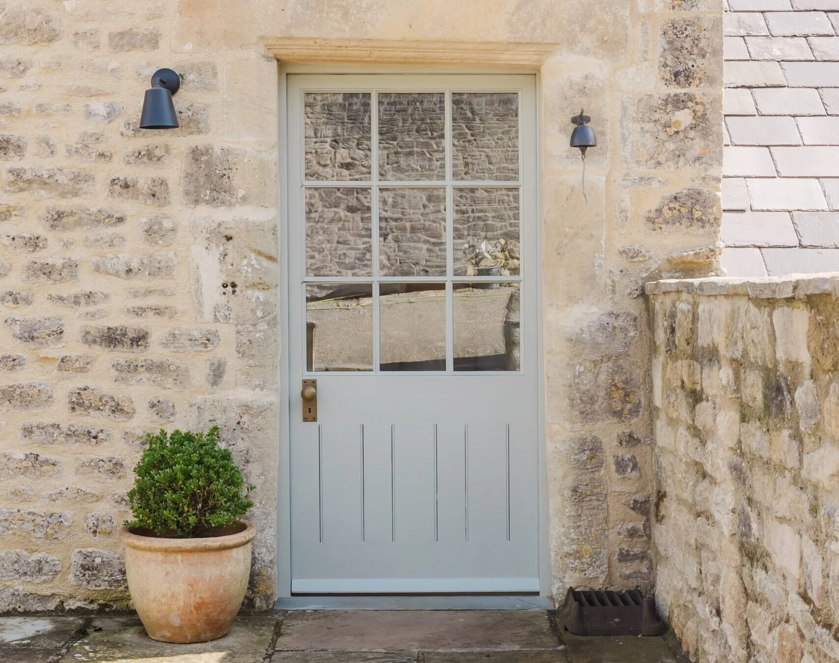 Exterior stone wall with a glass-paneled door, a potted plant at the left, and two black outdoor wall lights.