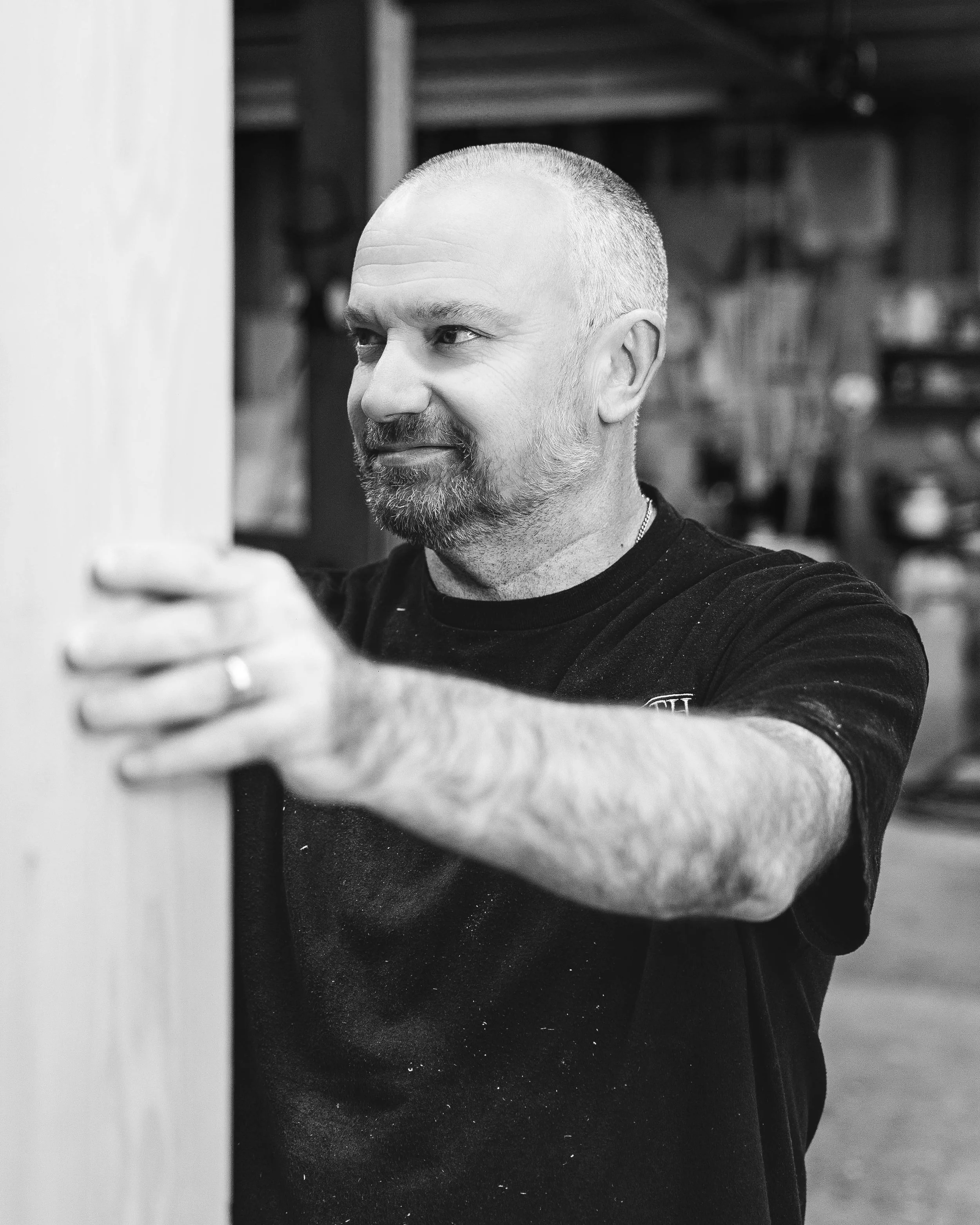 A man with a short beard and buzz cut wearing a black T-shirt, taking a selfie in a workshop or garage with shelves and various tools and objects in the background.
