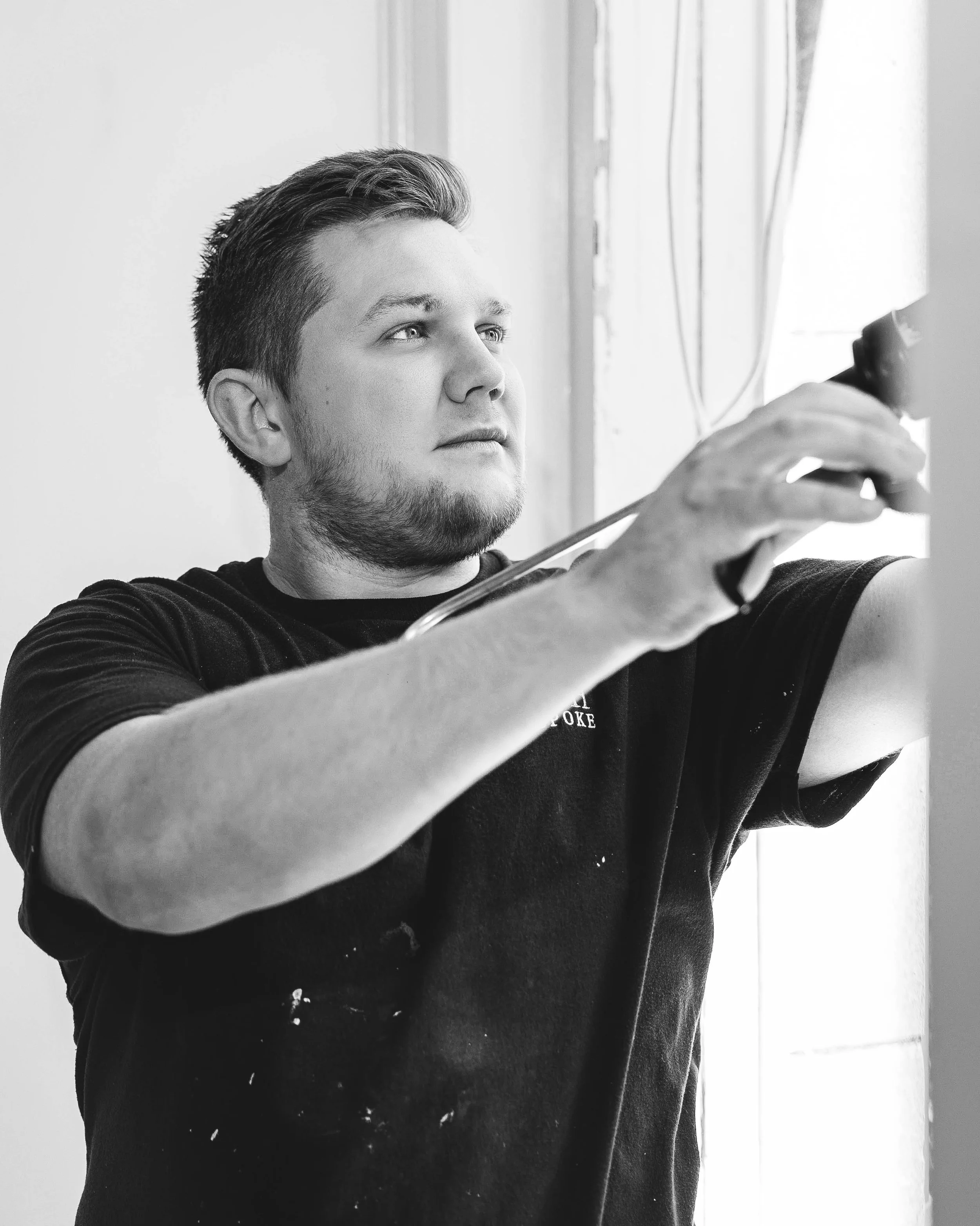 A black-and-white photo of a young man with styled hair and a beard, taking a selfie or adjusting a mirror, looking slightly to the side, in front of a window with curtains and some visible cords.