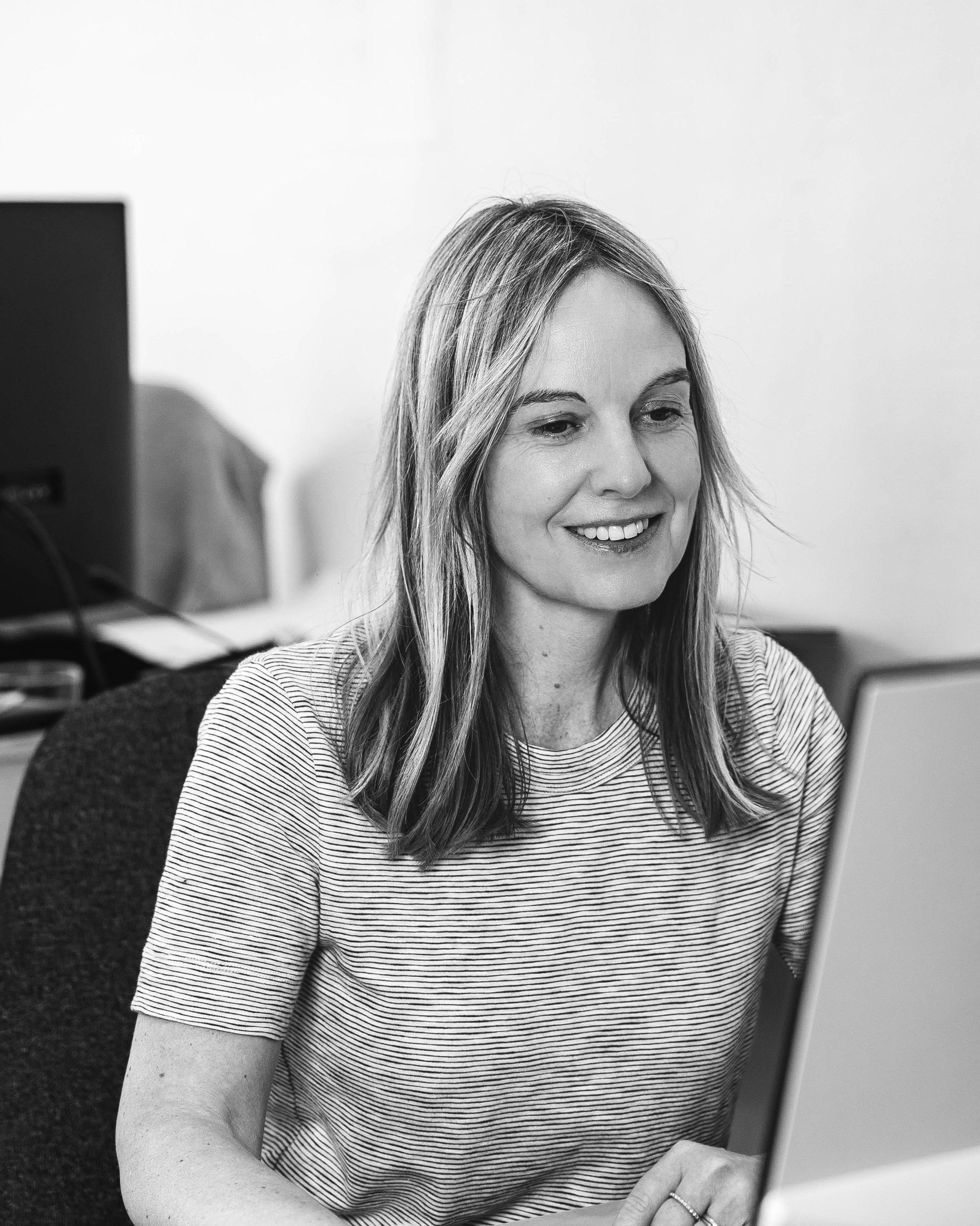 A woman with long hair is sitting at a desk, smiling, while looking at a computer screen.