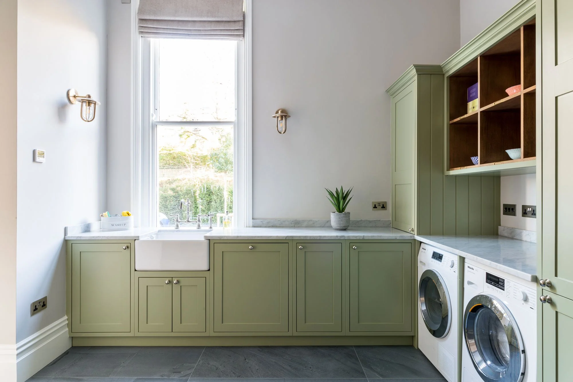 A kitchen with green cabinets, a white farmhouse sink, washing machines, a window with a roman shade, a small potted plant, and a marble countertop.