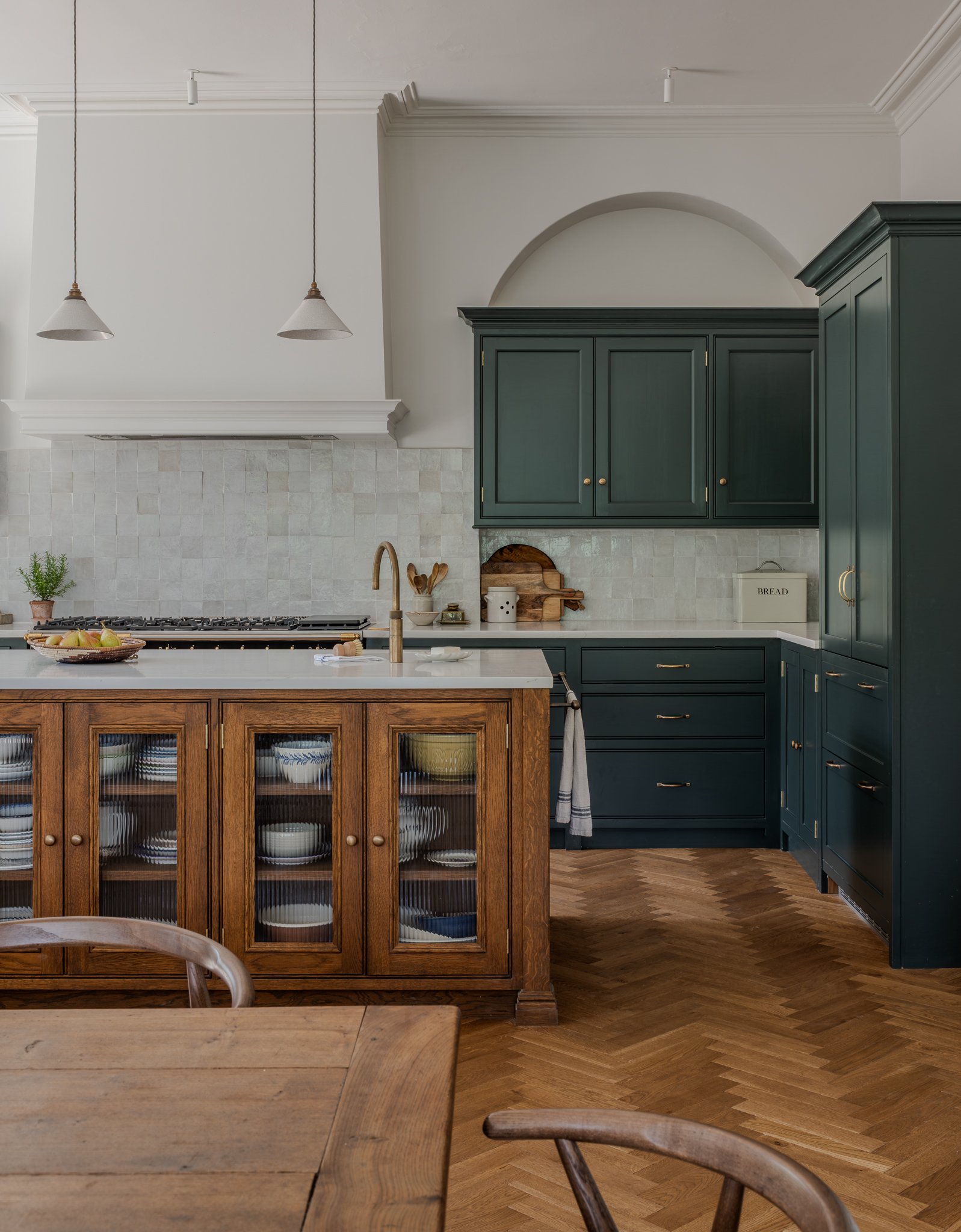 A kitchen with white upper walls, dark teal lower cabinets, a wooden kitchen island, a white tile backsplash, and wooden flooring. Two pendant lights hang from the ceiling, and there is a potted plant and a bowl of apples on the island.