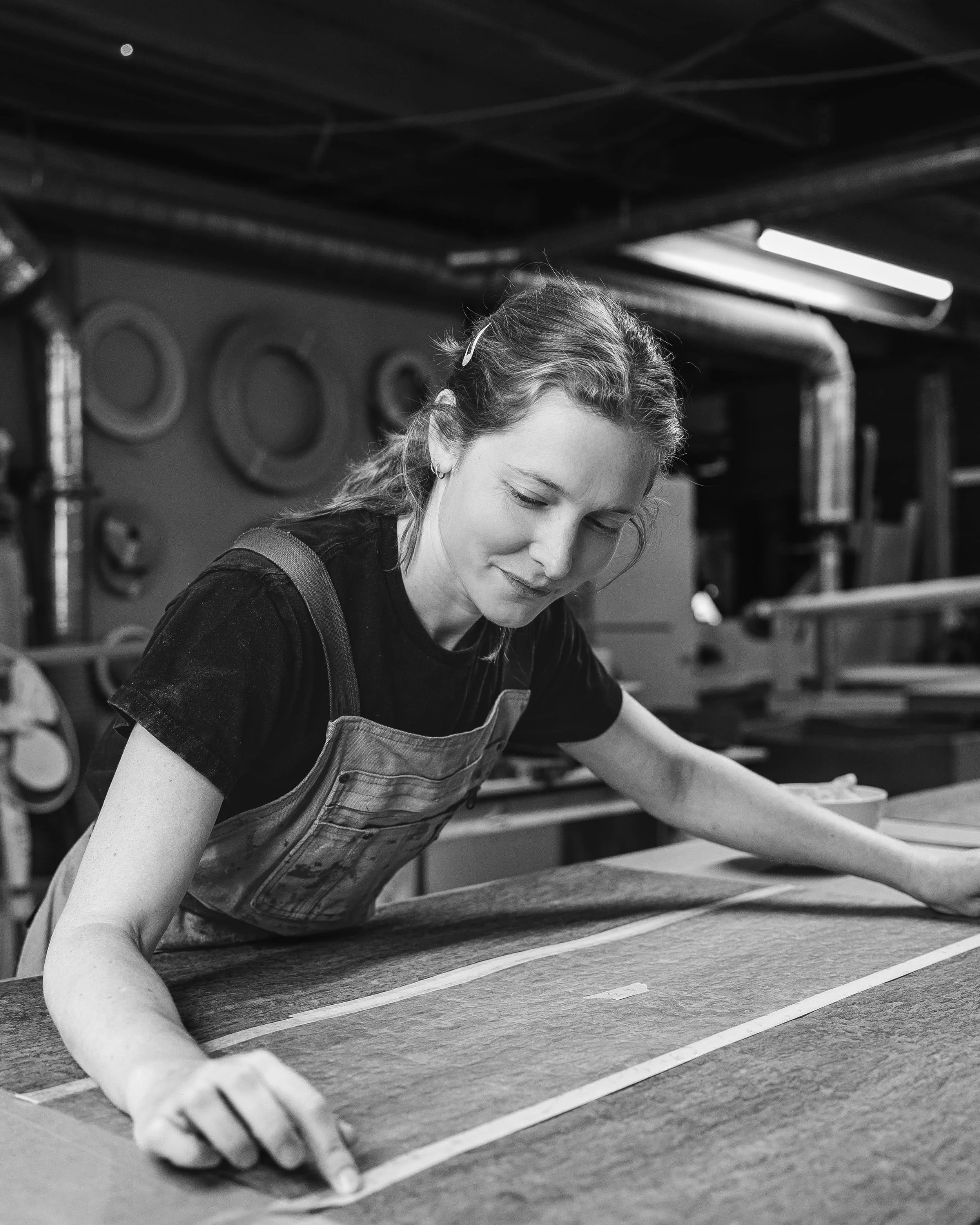 A woman working in a workshop, marking measurements on a piece of wood using a ruler and pencil, with tools and equipment in the background.
