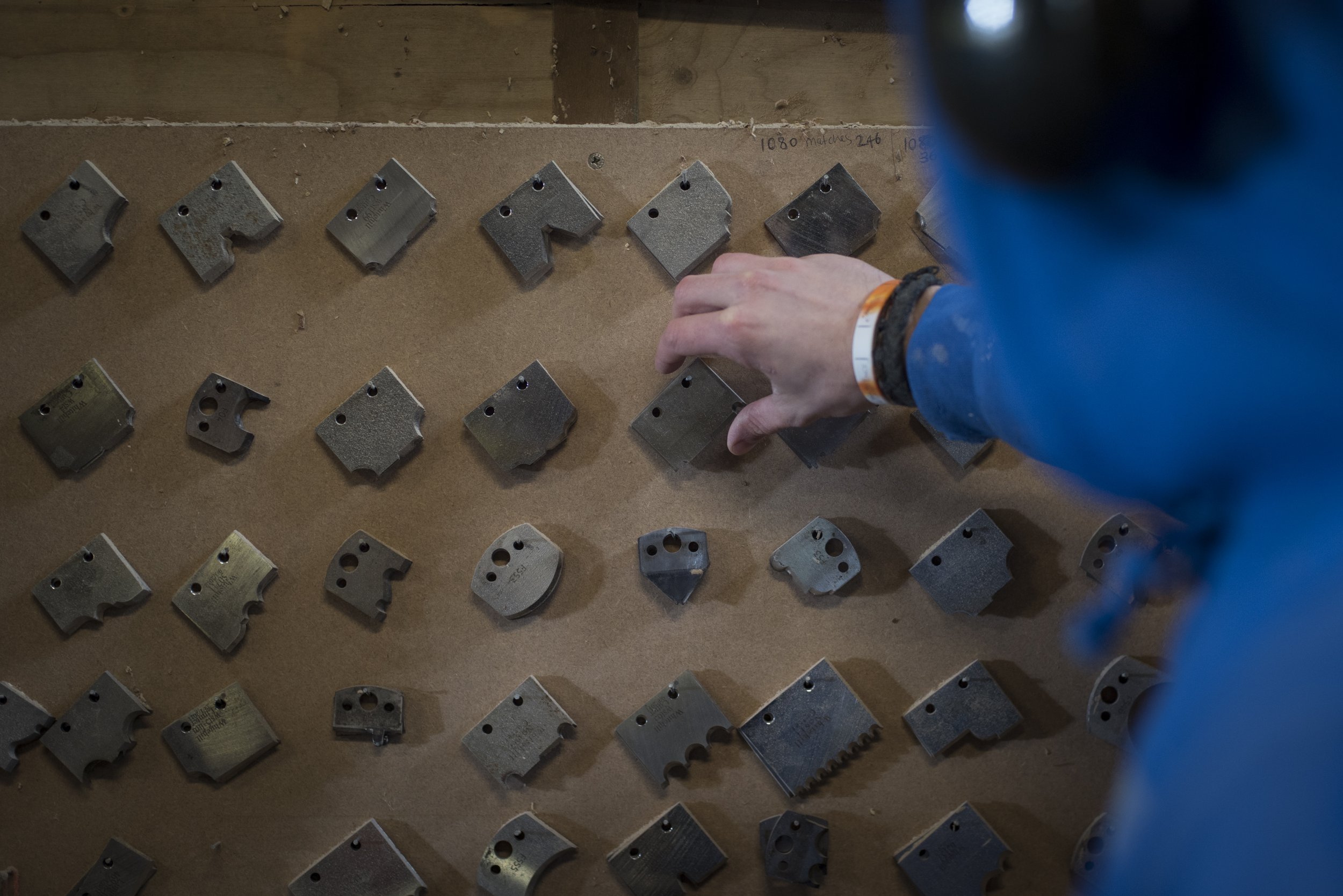 A person wearing a blue jacket and an orange wristband is placing small, gray metal brake pads on a wooden surface. There are multiple brake pads arranged in rows, along with some handwritten notes on the wood.