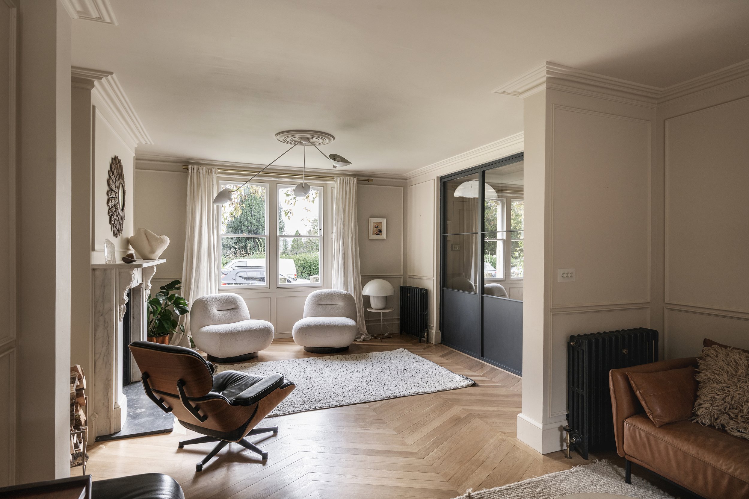 A living room with large windows, white curtains, beige walls with decorative molding, a white fireplace mantel with decorative items, white armchairs, a modern wood lounge chair, a black radiator, and a sliding glass partition separating another roo