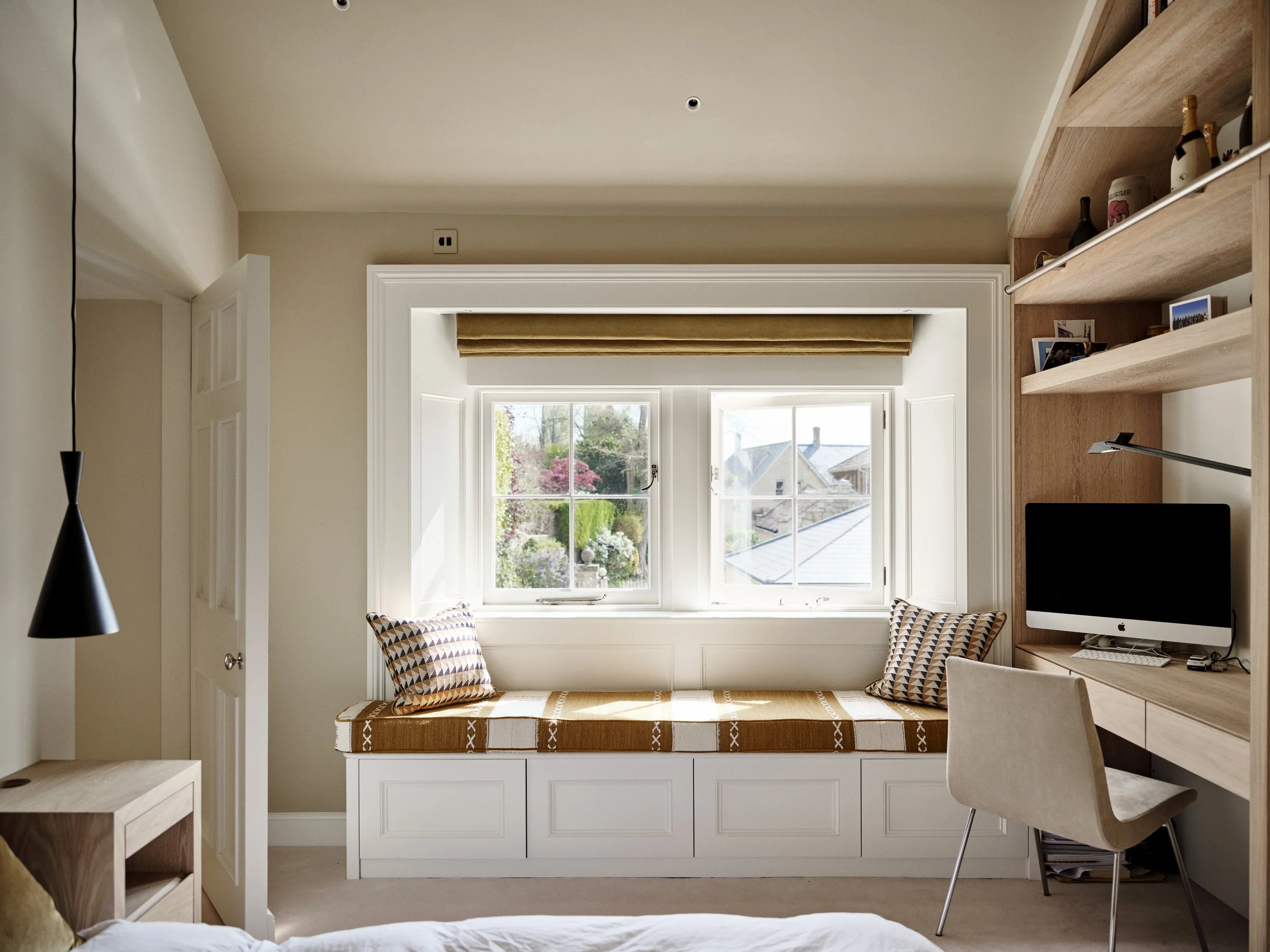 A cozy bedroom with a large window letting in natural light. There is a window seat with patterned cushions below the window, a wooden desk with a computer, and built-in shelves holding decorative items and books. The room has beige walls and a white ceiling.