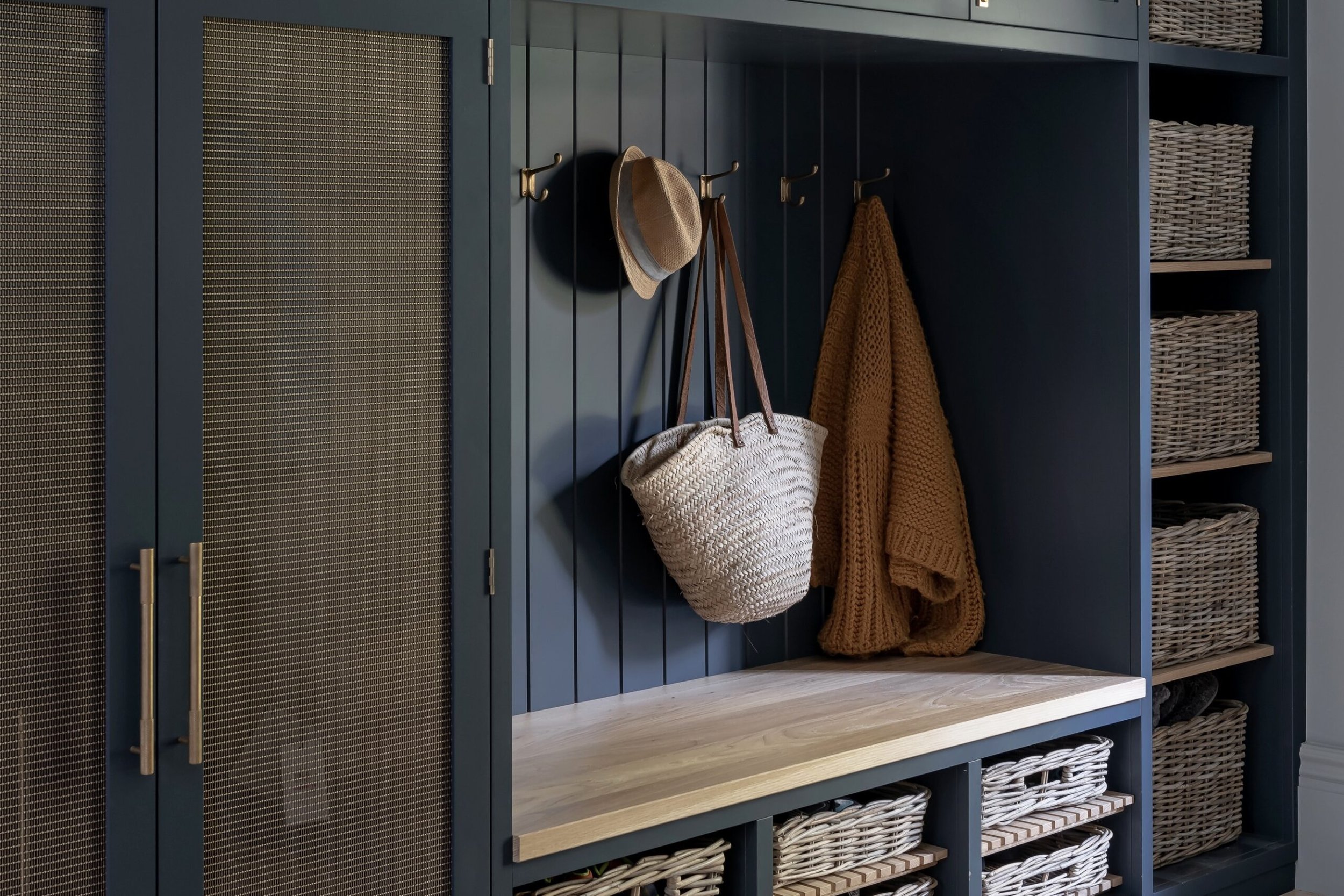 A navy blue storage cabinet with hooks holding a beige hat, brown tote bag, and orange knit blanket. The cabinet has woven baskets on open shelves and a light wood bench at the bottom.