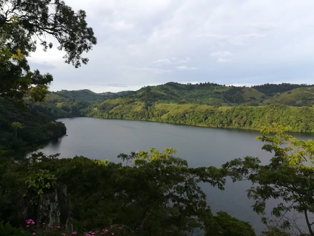 A scenic view of a large body of water, surrounded by rolling green hills and trees, with cloudy skies overhead.