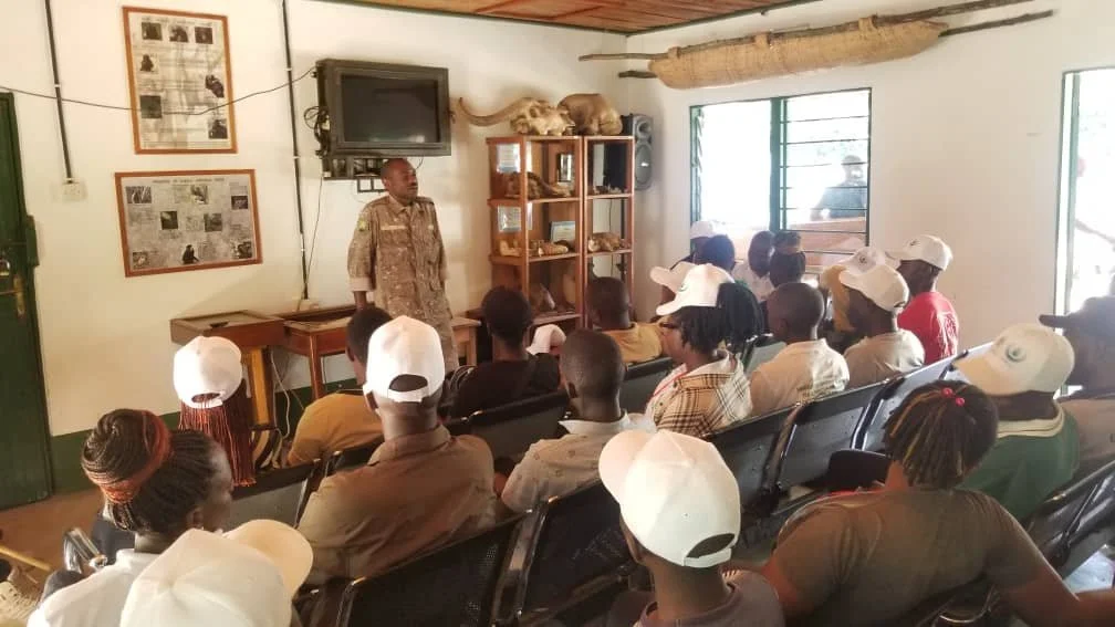 A man in military camouflage uniform giving a presentation to a group of people sitting in a classroom or meeting room. The room contains shelves with animal skulls and bones, and has windows letting in natural light.