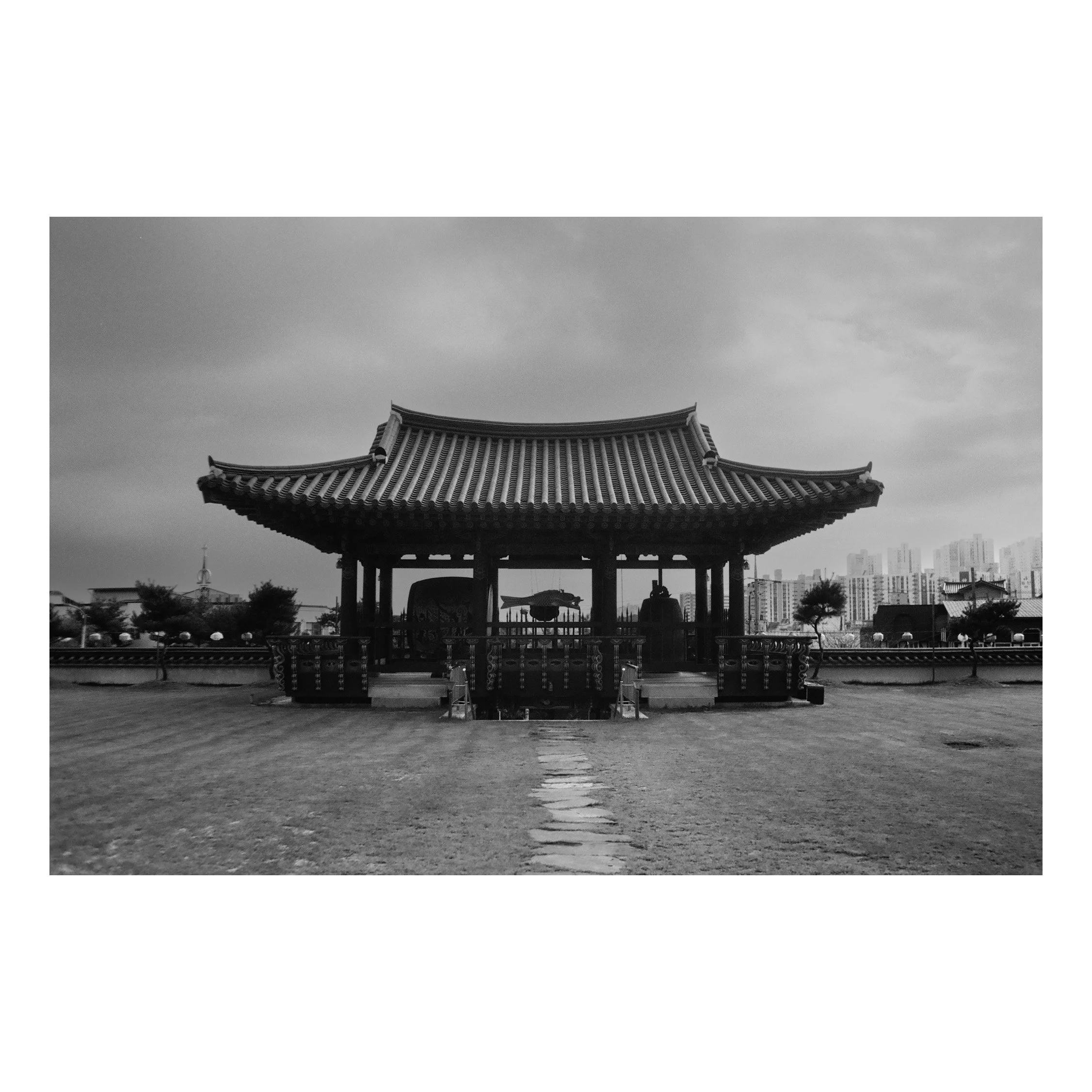Traditional Korean pavilion with ornate roof and statues inside, trees and modern buildings in the background, black and white photo.